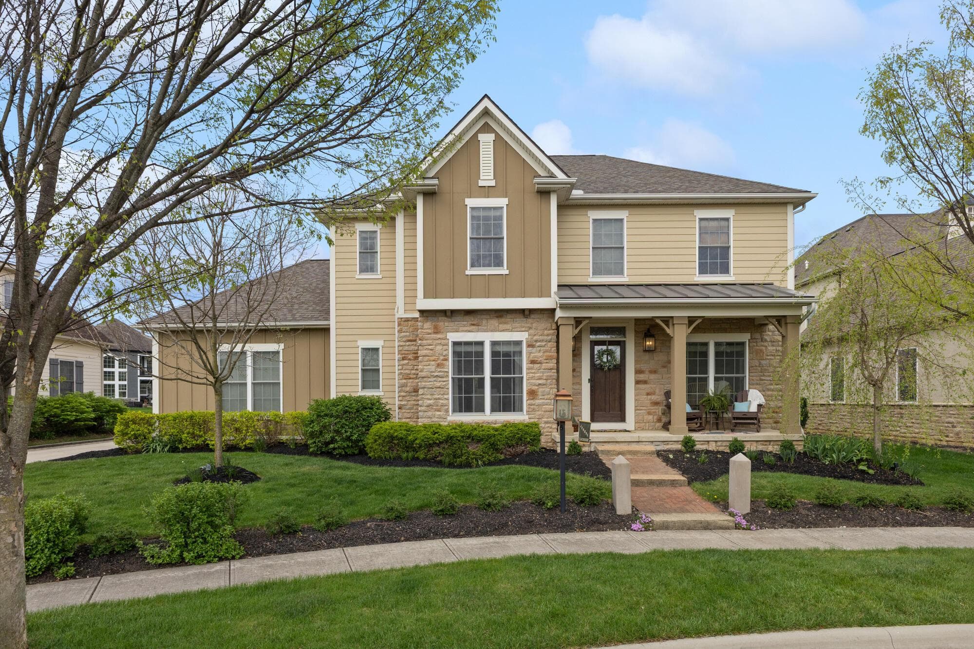 Tan and stone two-story home with a covered front porch, brick front walk, mature landscaping, and a well-maintained lawn.