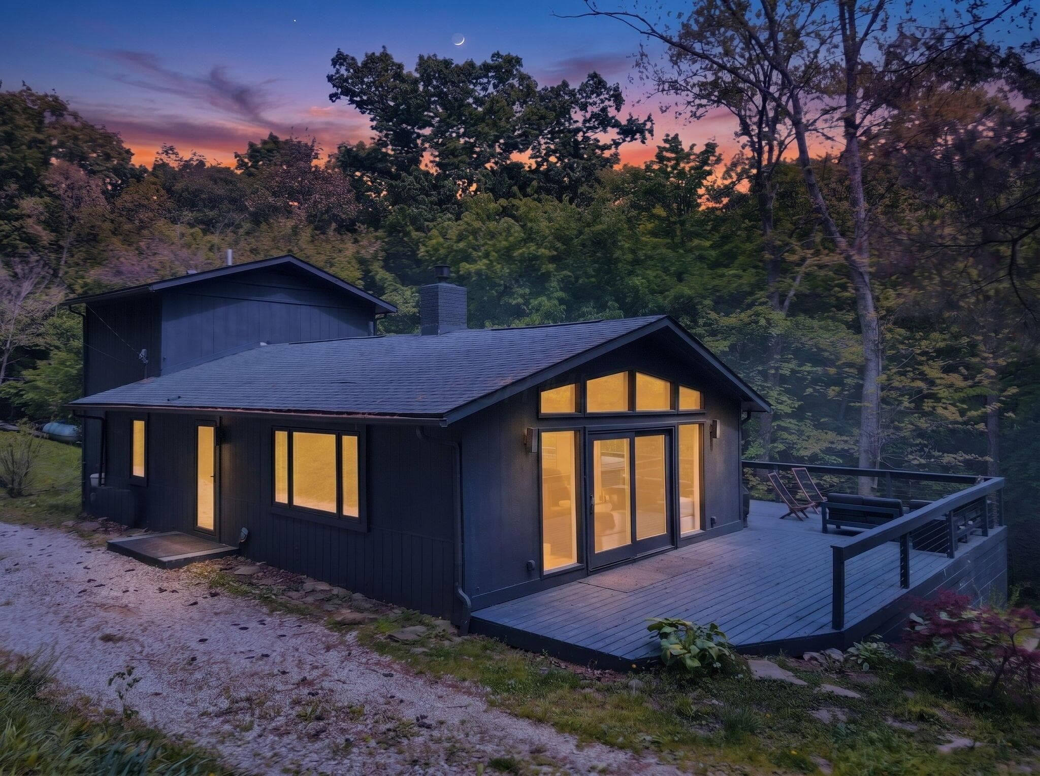 Modern cabin retreat surrounded by mature trees, featuring a spacious wraparound deck, expansive windows, and warm interior glow that creates an inviting atmosphere from dusk to dawn.