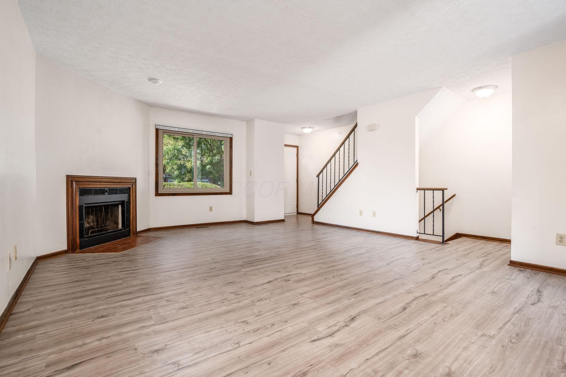 Open living area featuring LVT flooring, a corner fireplace with a wood surround, a window with exterior views, and a staircase with dark wood handrails and black metal spindles