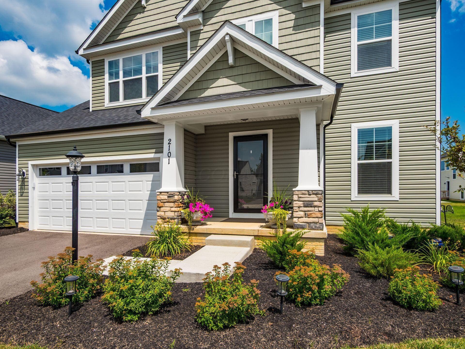 Inviting front facade featuring a covered entry with stone-accented columns, light green siding, and a prominent white garage door