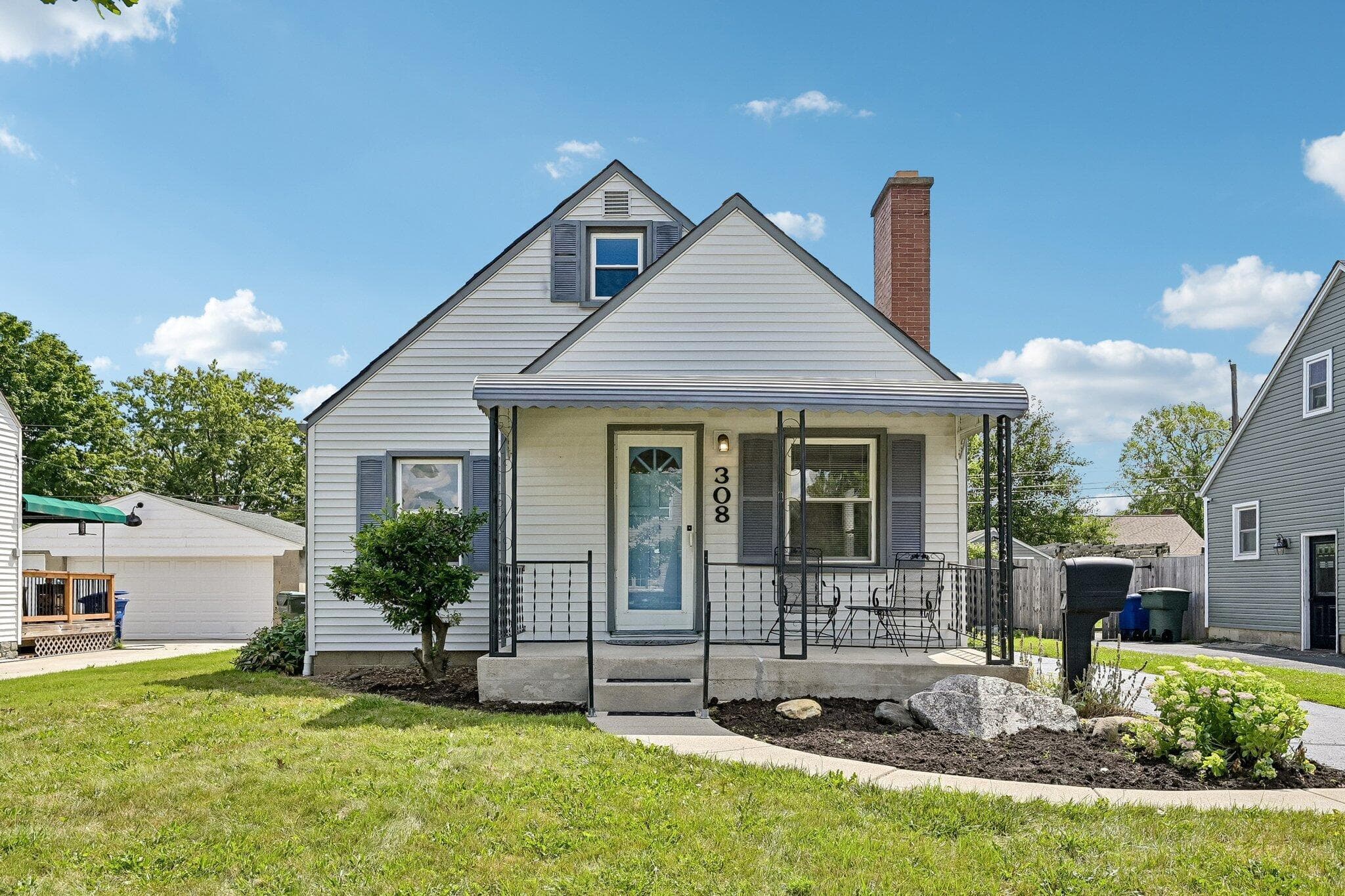 Light-colored siding exterior with dark gray shutters, a covered front porch featuring wrought iron railing, a brick chimney, and an attached single-car garage
