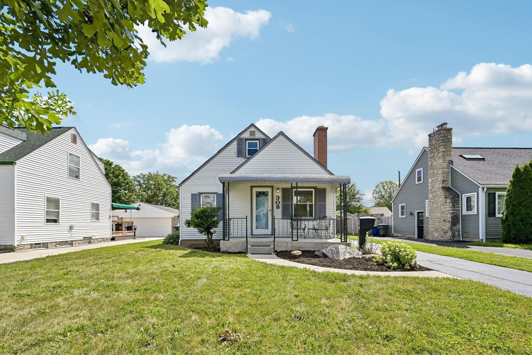 White siding residence featuring a covered front porch with metal railings, a brick chimney, and a landscaped front yard with a rock feature