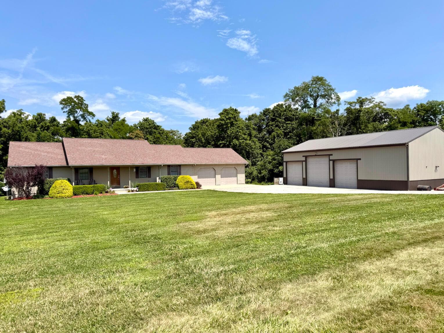 Expansive grounds featuring a main residence with a two-car garage, a separate large metal outbuilding with three bay doors, a paved driveway, mature trees, and landscaping