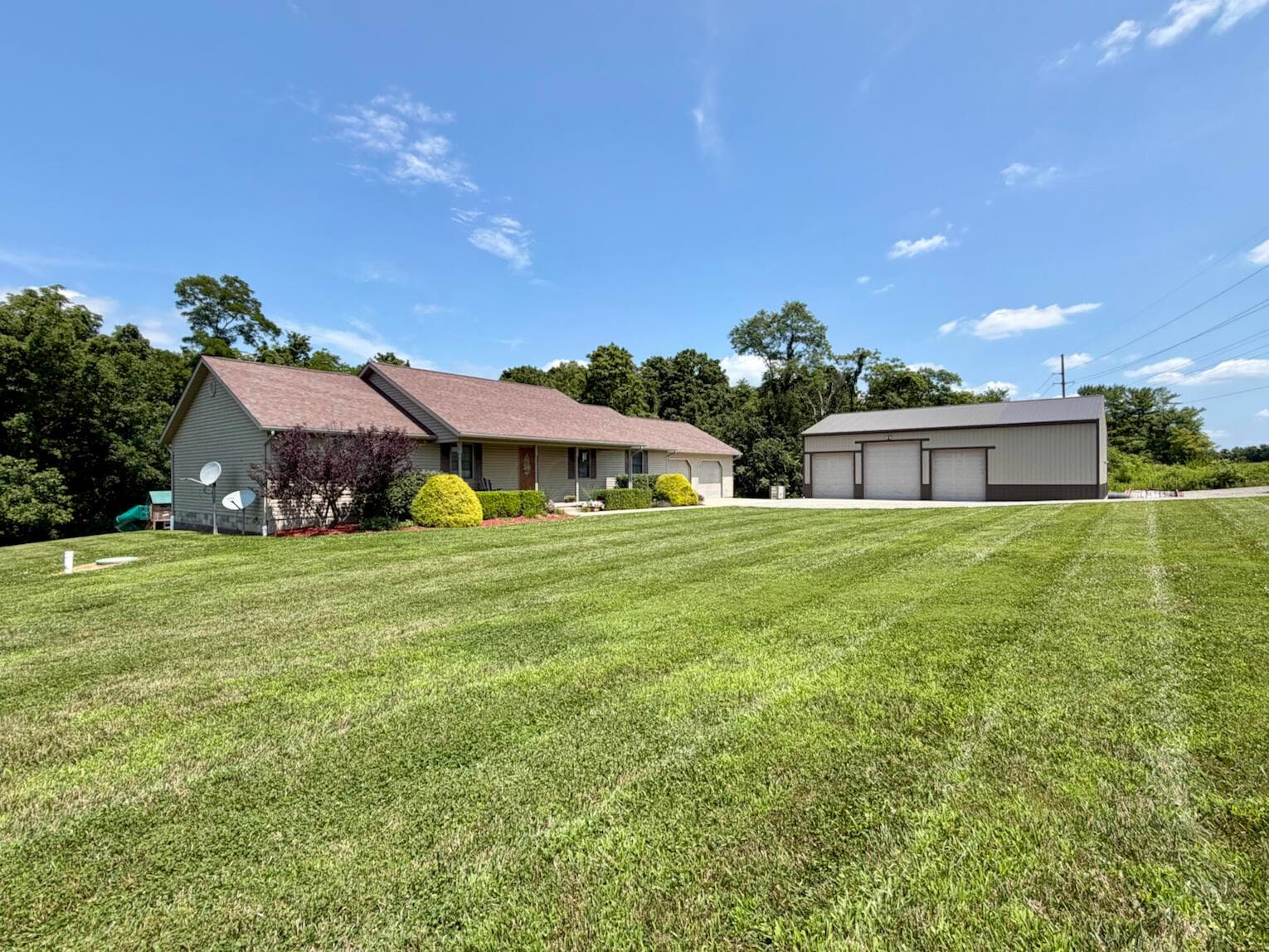Expansive green lawn presenting a siding-clad home with a brown shingle roof and an additional multi-bay garage structure