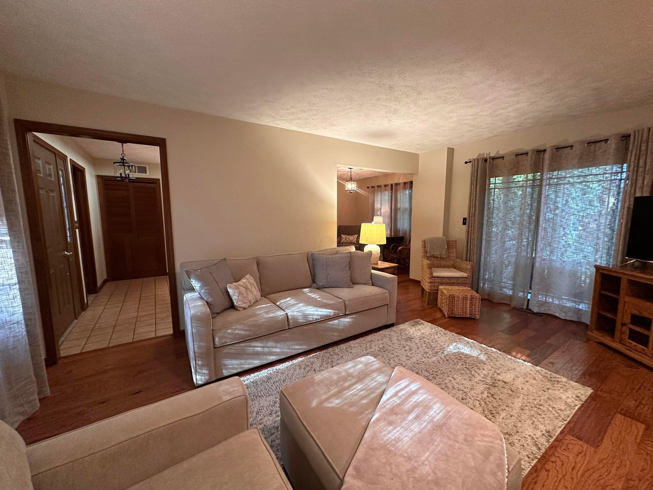 Spacious living area featuring wood-finish flooring, light-toned walls, and a large window with sheer drapes