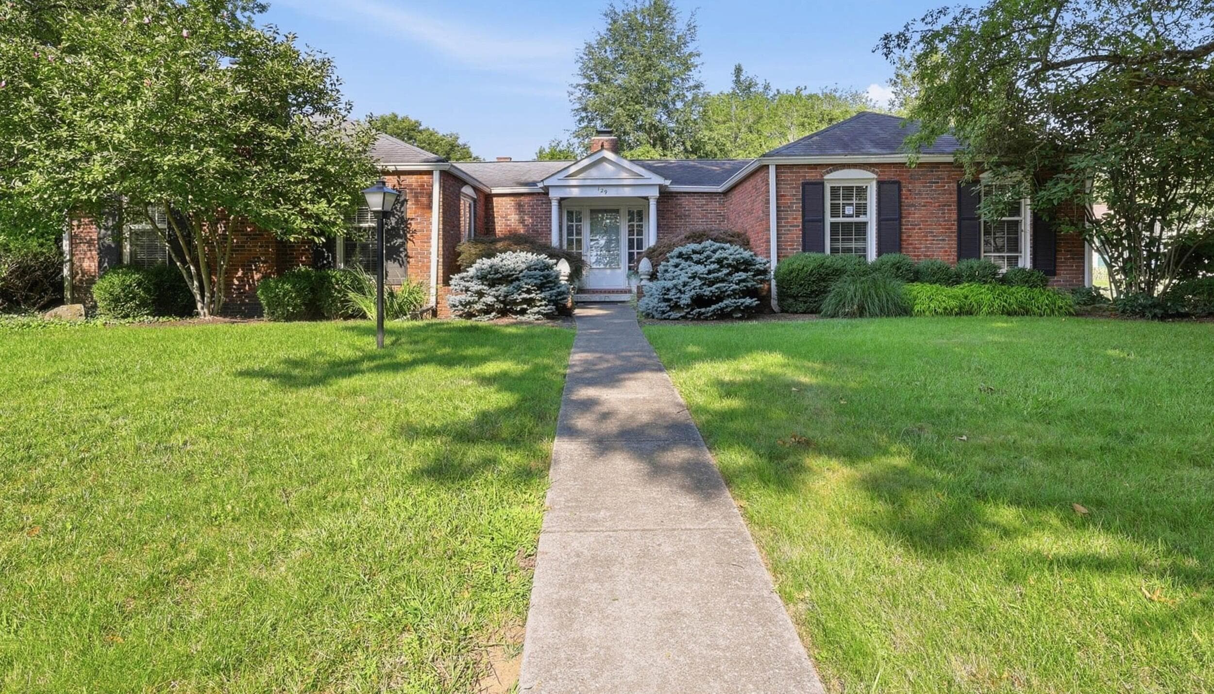 Brick exterior featuring a gabled entry, prominent windows with dark shutters, established landscaping, and a concrete pathway leading to the front door