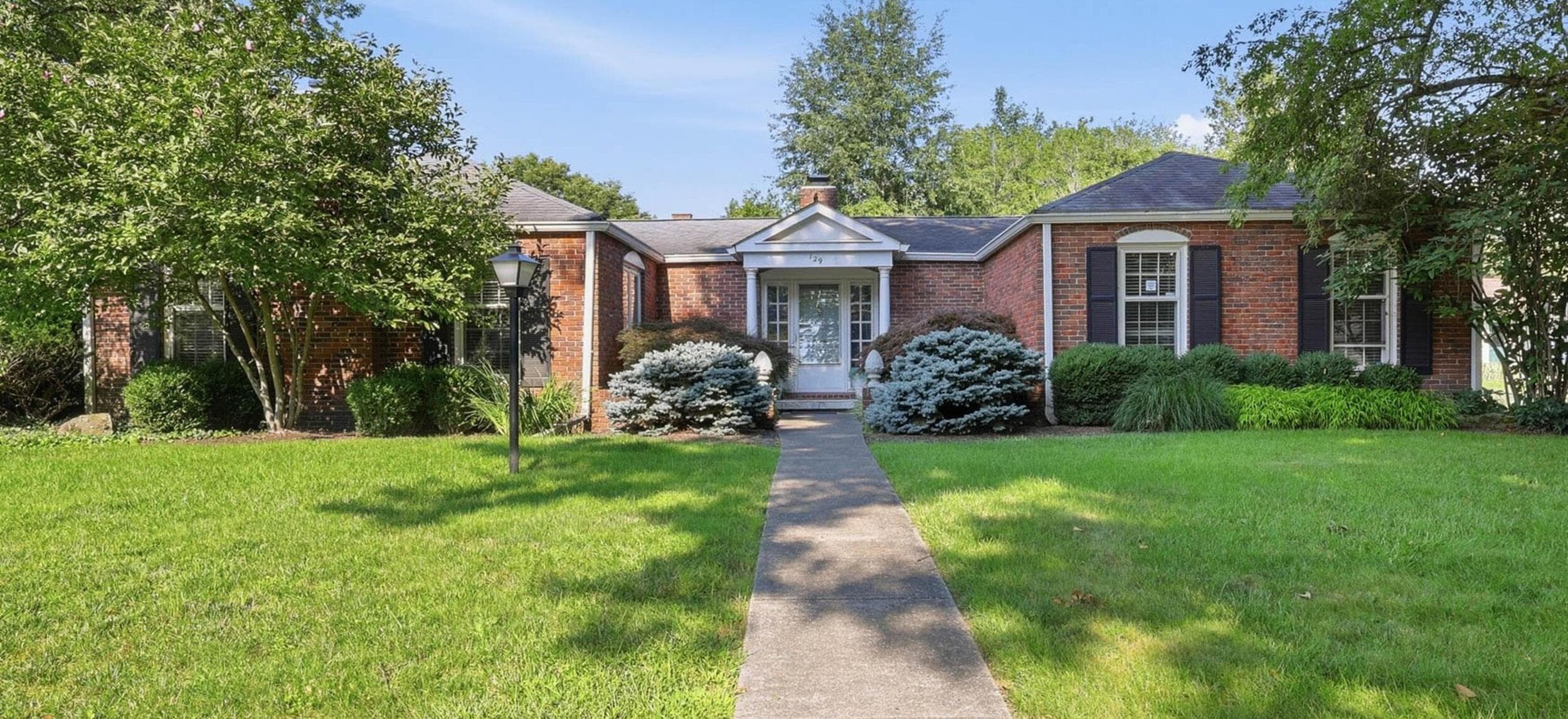 Brick exterior featuring a gabled entry, prominent windows with dark shutters, established landscaping, and a concrete pathway leading to the front door