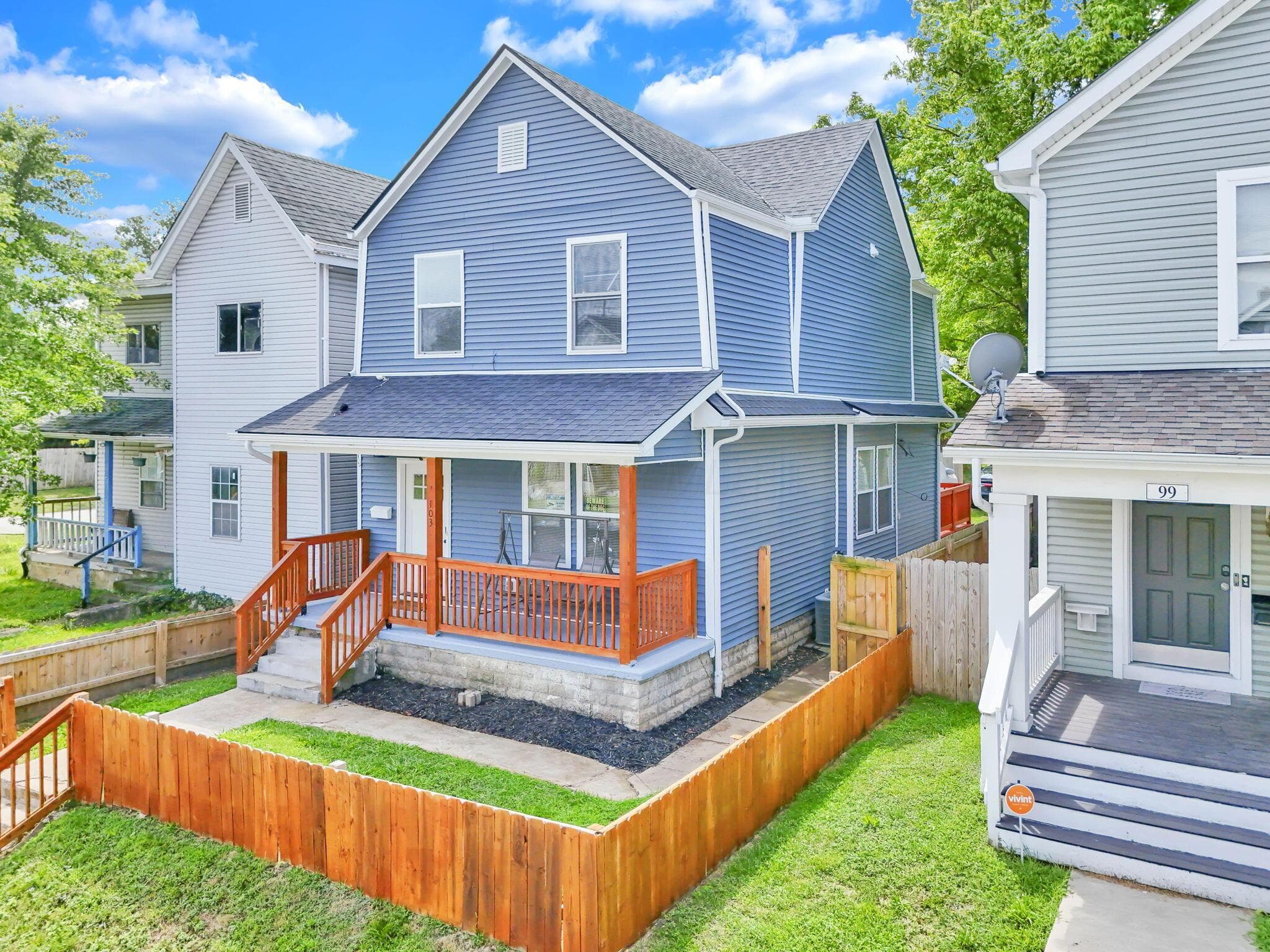 Two-story residence featuring blue siding, a covered front porch with natural wood railings, a dark shingle roof, and a fully fenced front yard