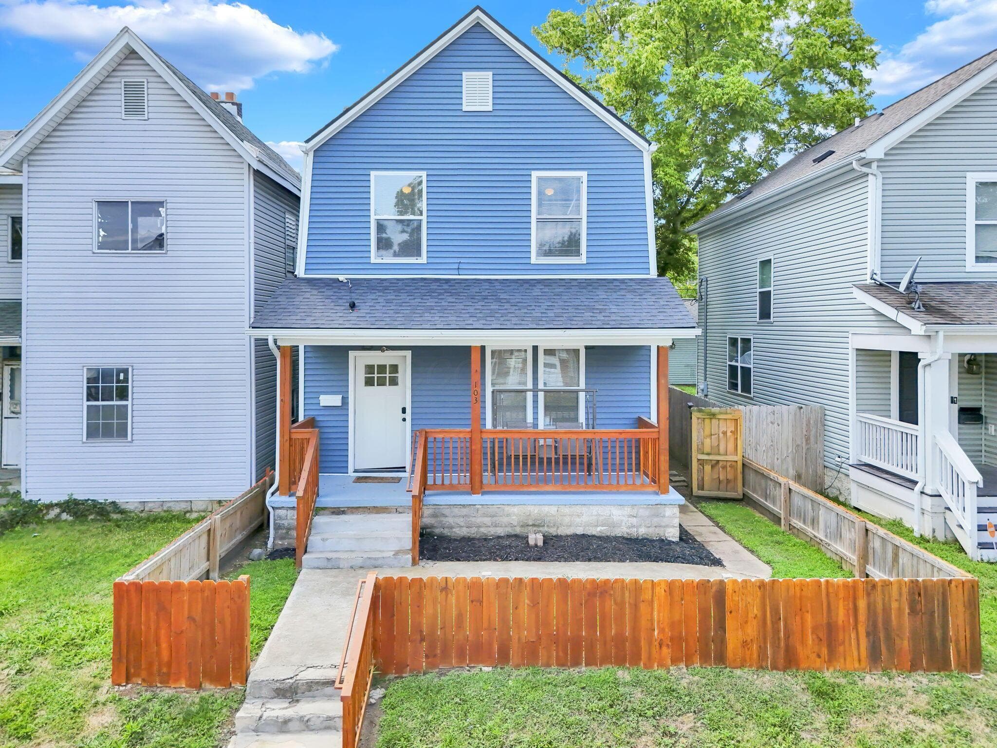 Blue siding exterior featuring a covered front porch with wood railings, a white entry door, and a partially fenced front yard