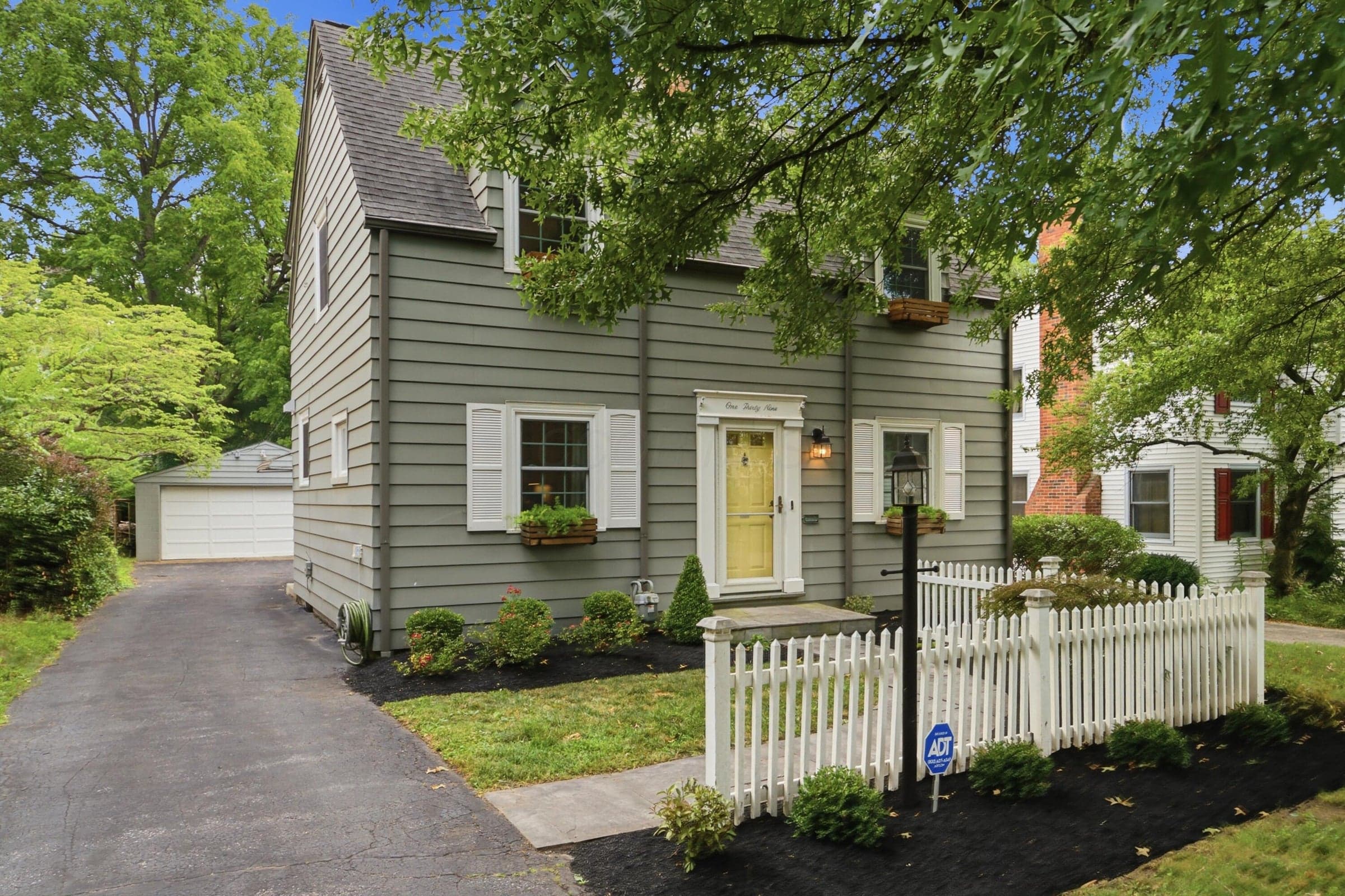 Adorable Clintonville home featuring a gray siding exterior, white picket fence, paved driveway, detached garage, and mature trees