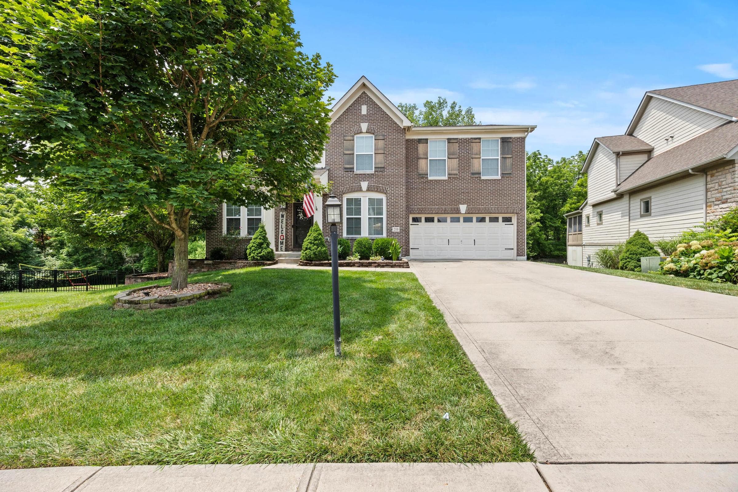 Brick facade residence featuring shuttered windows, an attached two-car garage, a concrete driveway, and a landscaped front yard with mature trees