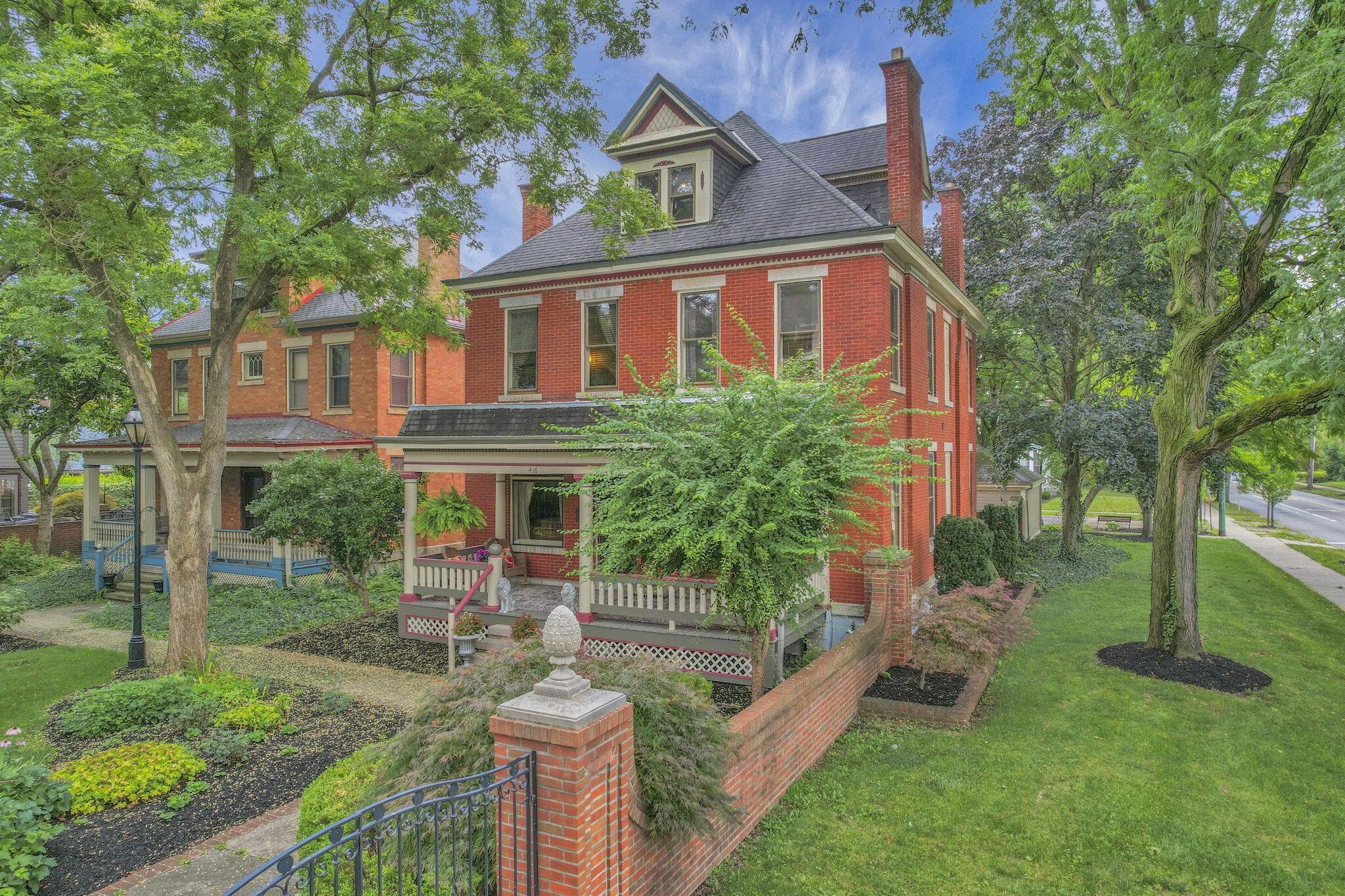 Brick exterior with multiple chimneys, covered front porch, dormer window, and mature landscaping