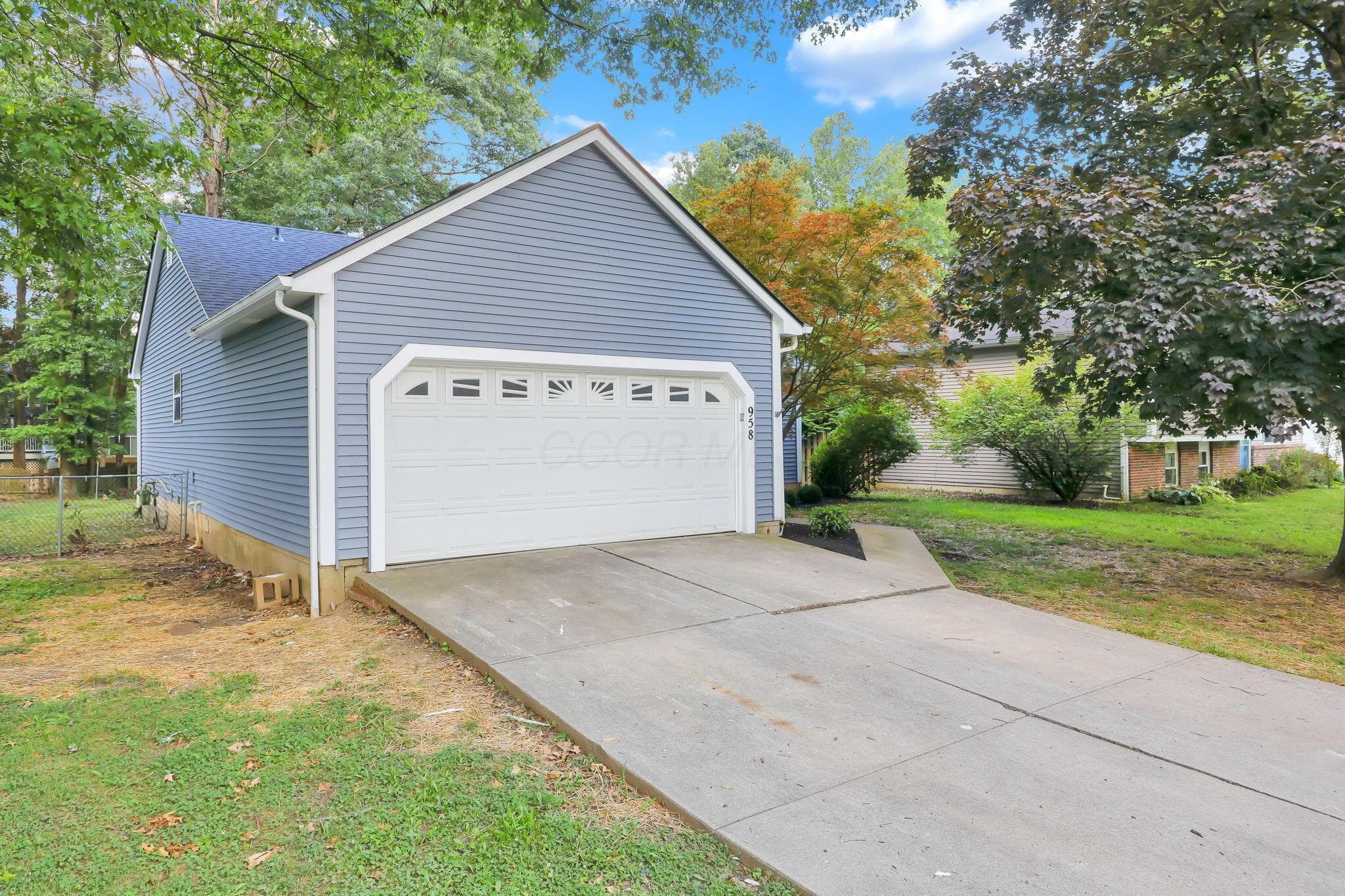 Attached two-car garage with white paneled door, blue vinyl siding, concrete driveway, and established surrounding trees