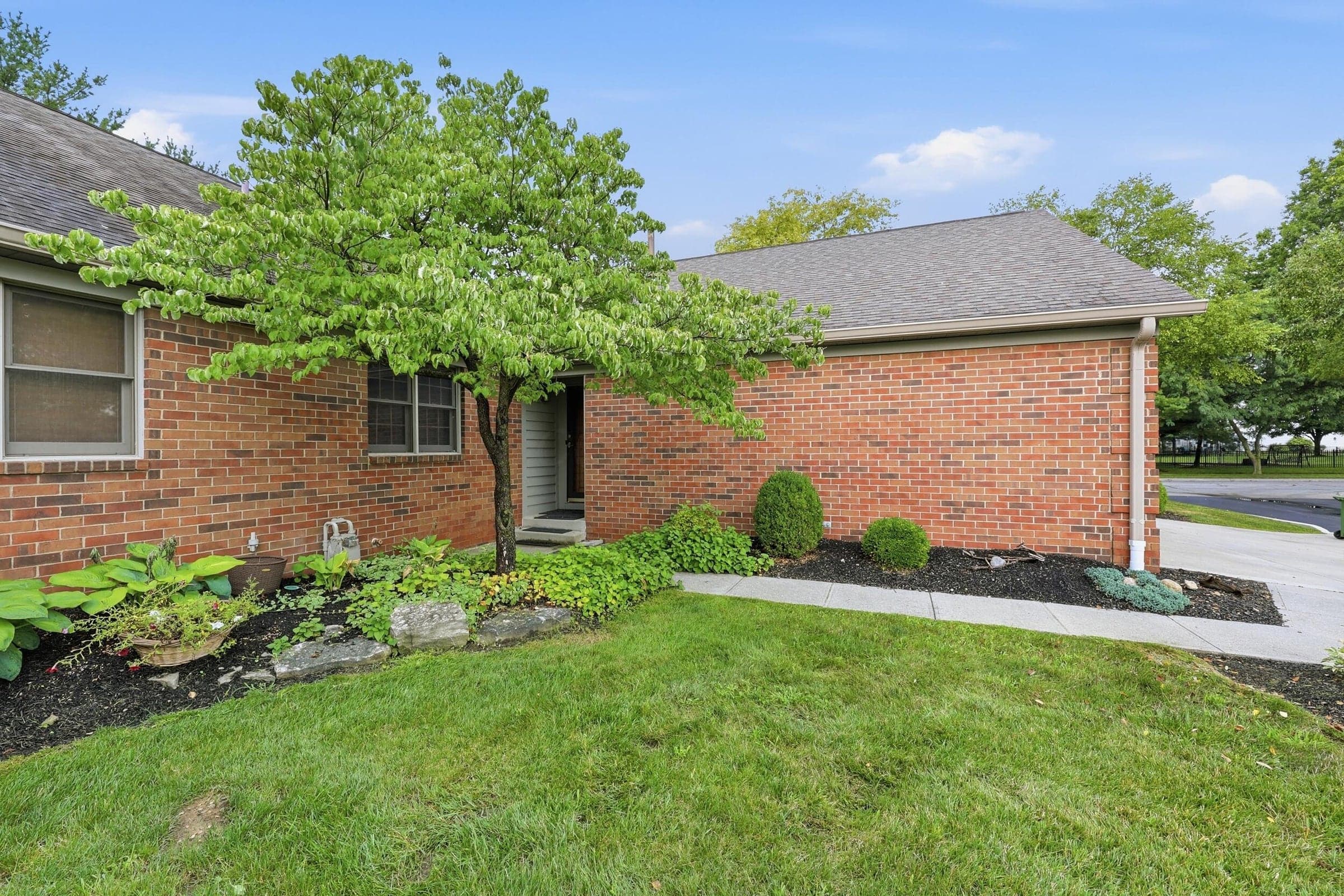 Brick exterior with a mature tree, landscaped garden beds, a concrete walkway, and a neatly maintained lawn