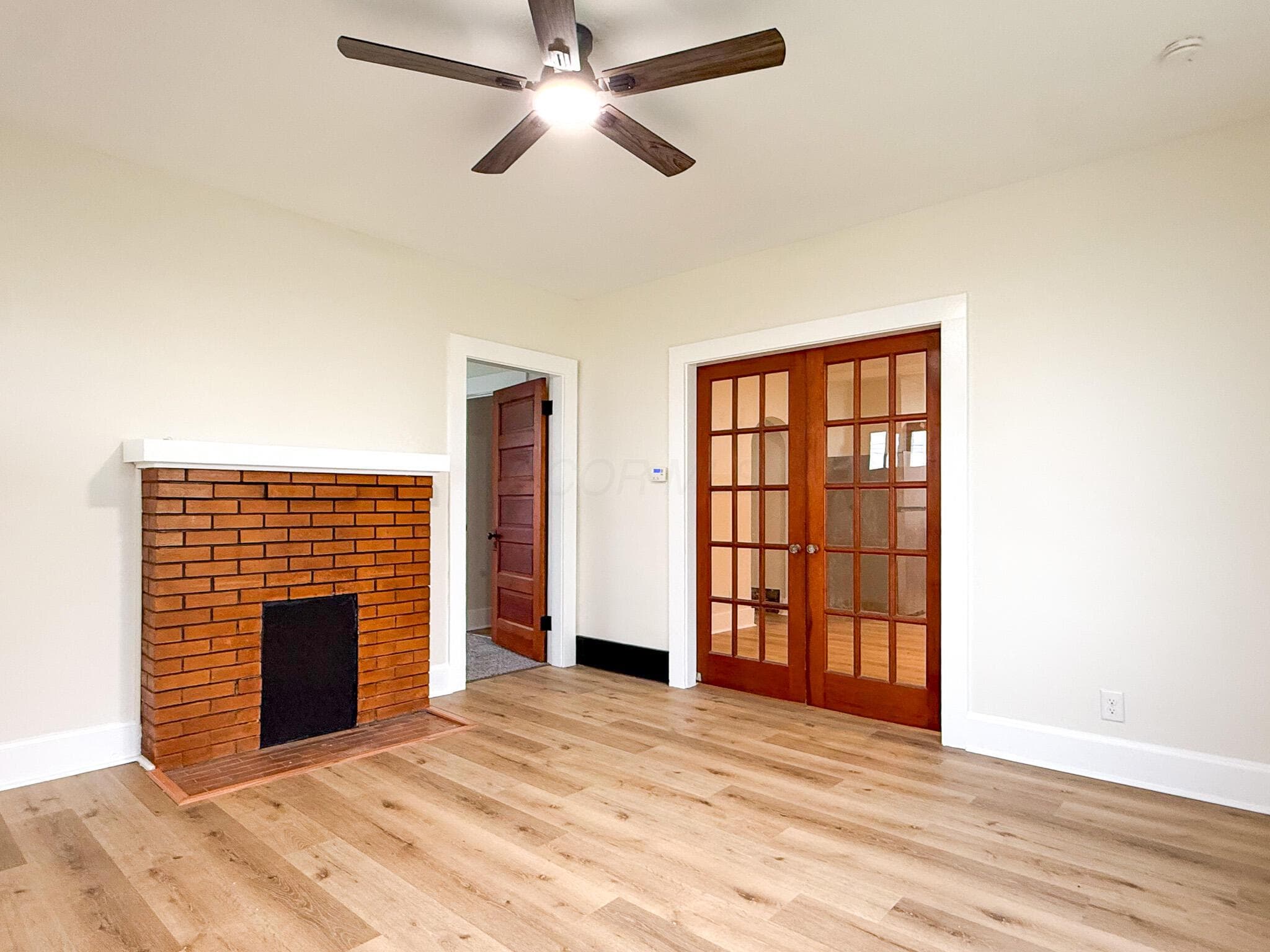 Wood-finish flooring, brick fireplace with a white mantel, wood-framed double French doors, and a ceiling fan with integrated lighting
