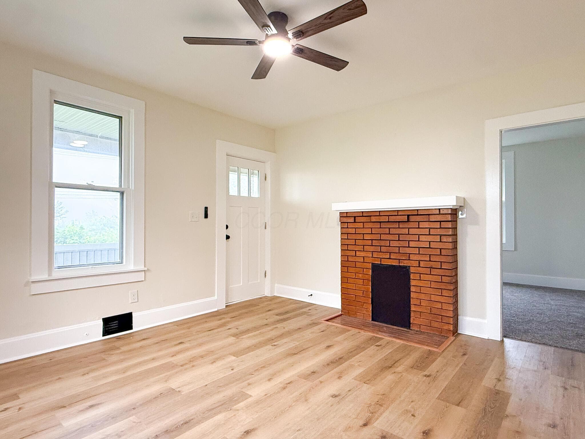 Wood-finish flooring, brick fireplace with white mantel, white trim, ceiling fan, and white paneled door with glass inserts