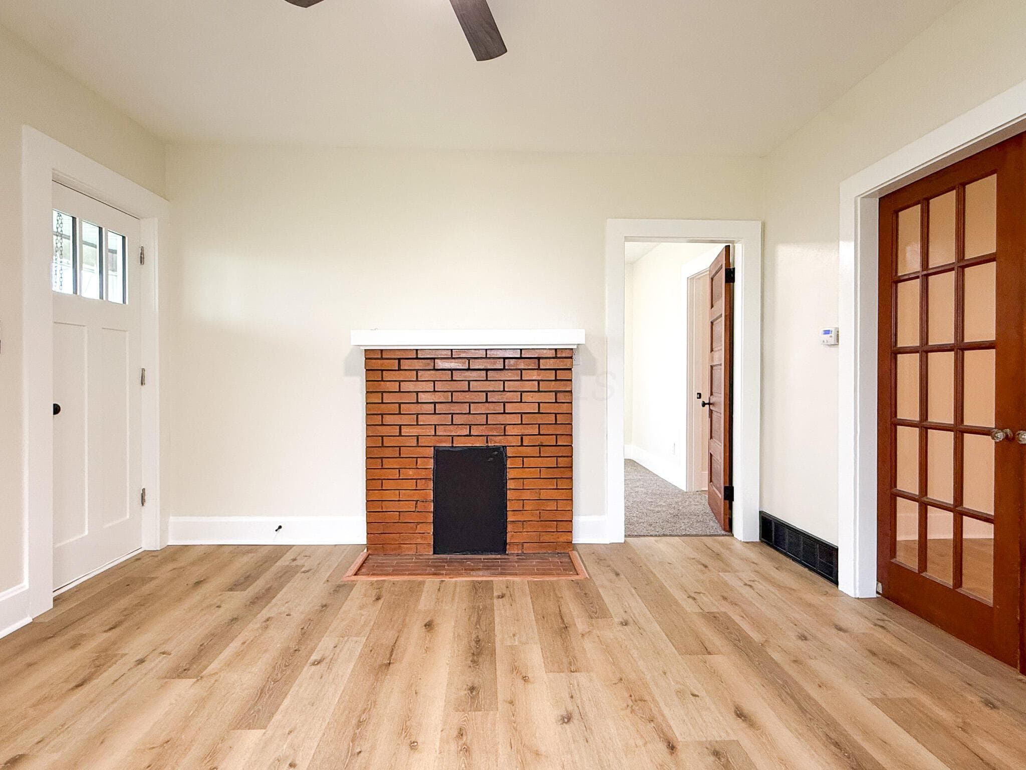 Wood-finish flooring, brick fireplace with a white mantel, white trim, white interior door with glass panels, and a dark wood interior door with glass panels