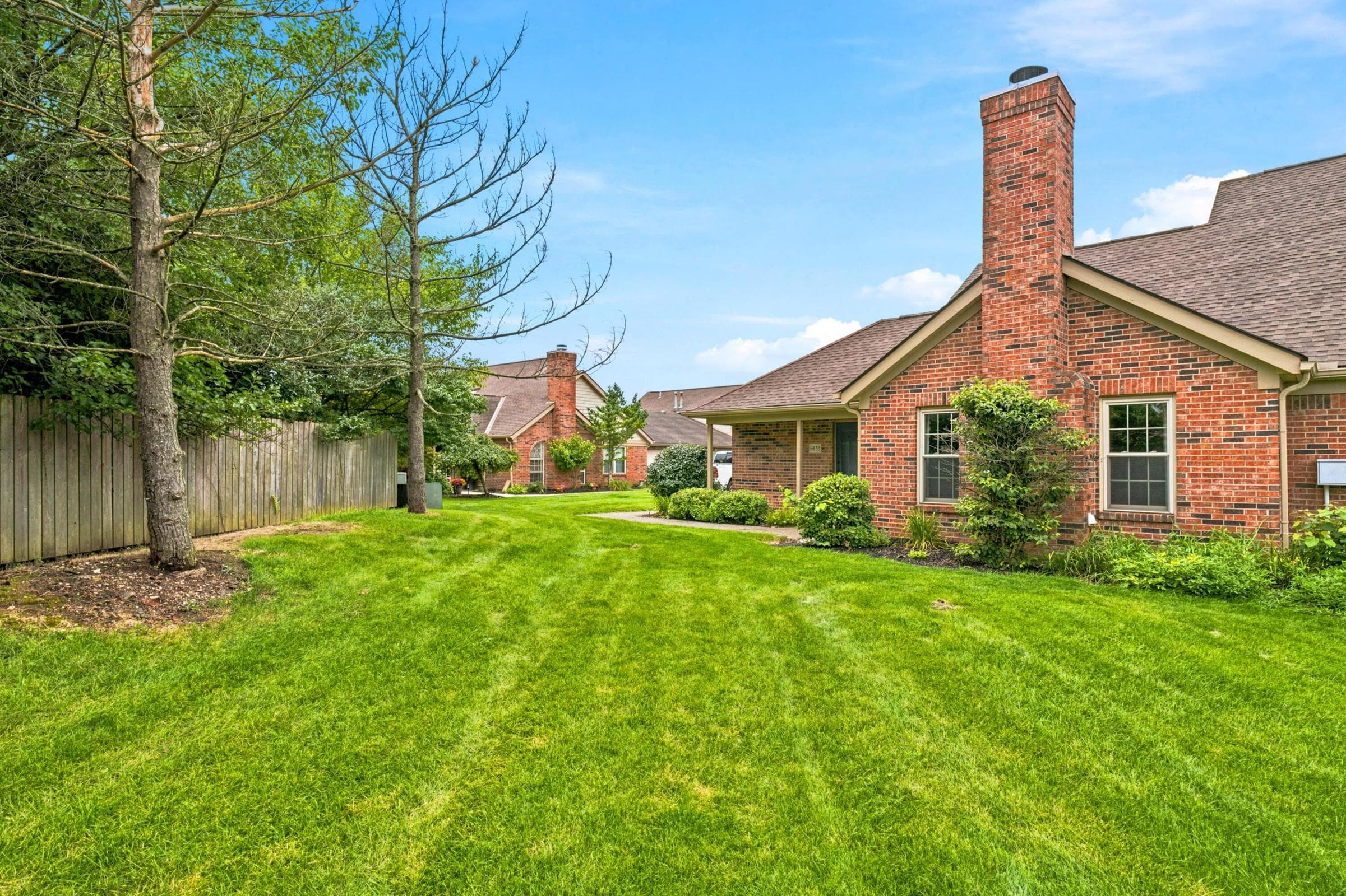 Expansive green lawn with a wooden fence, surrounding landscaping, and a brick exterior with a prominent chimney