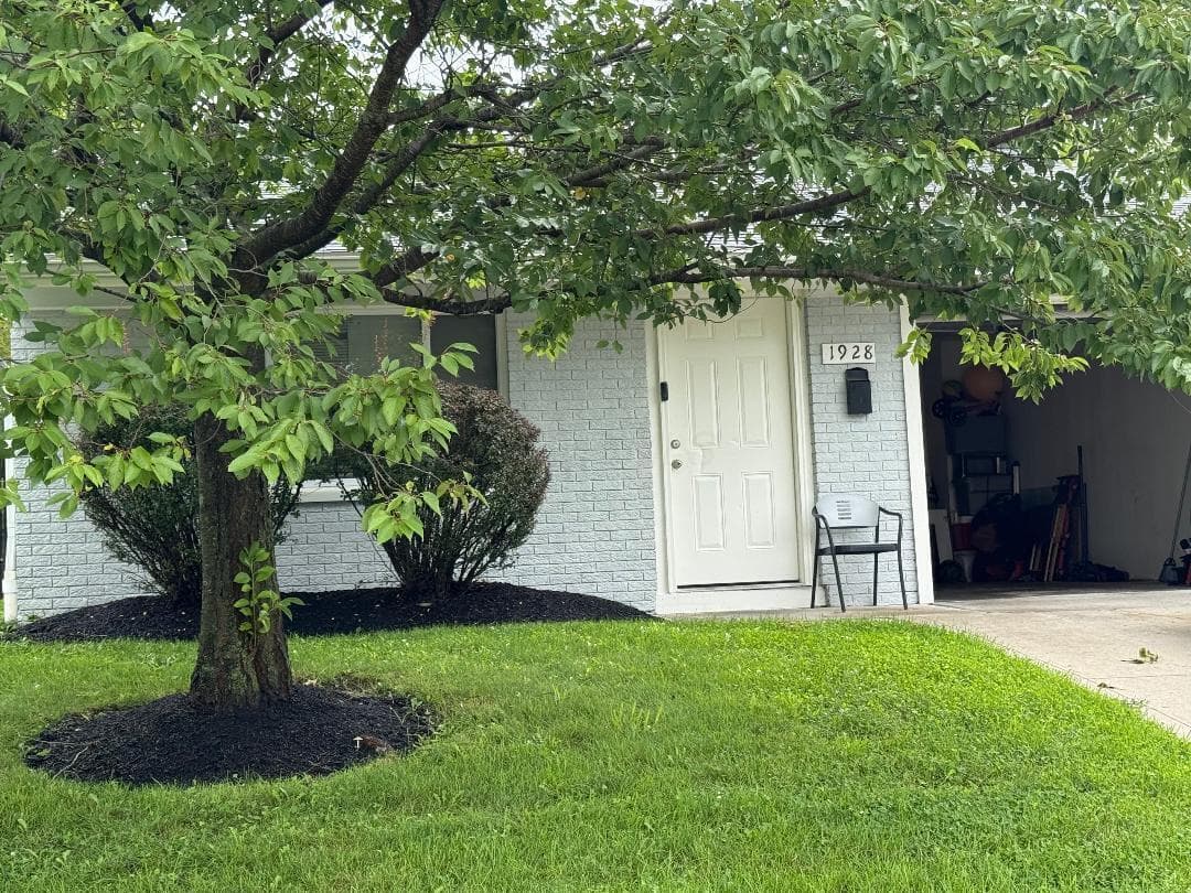 Painted brick exterior with a white entry door, an attached garage, and a manicured lawn with mature trees