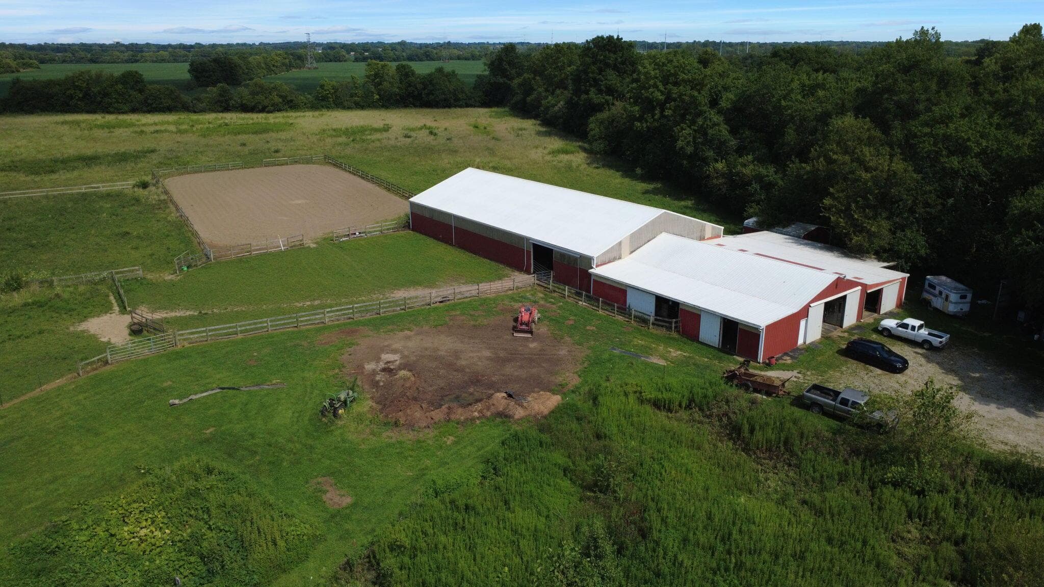 Red-sided barn structures with white roofs, a fenced riding arena, and expansive green acreage