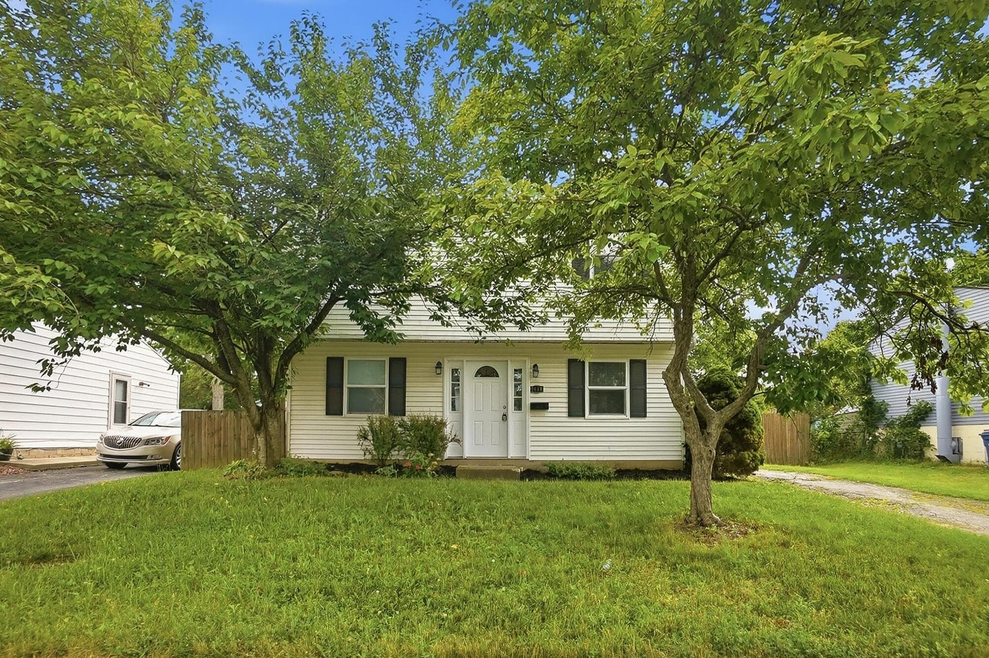 White vinyl siding exterior with dark shutters, front entry door, and mature trees in the front yard