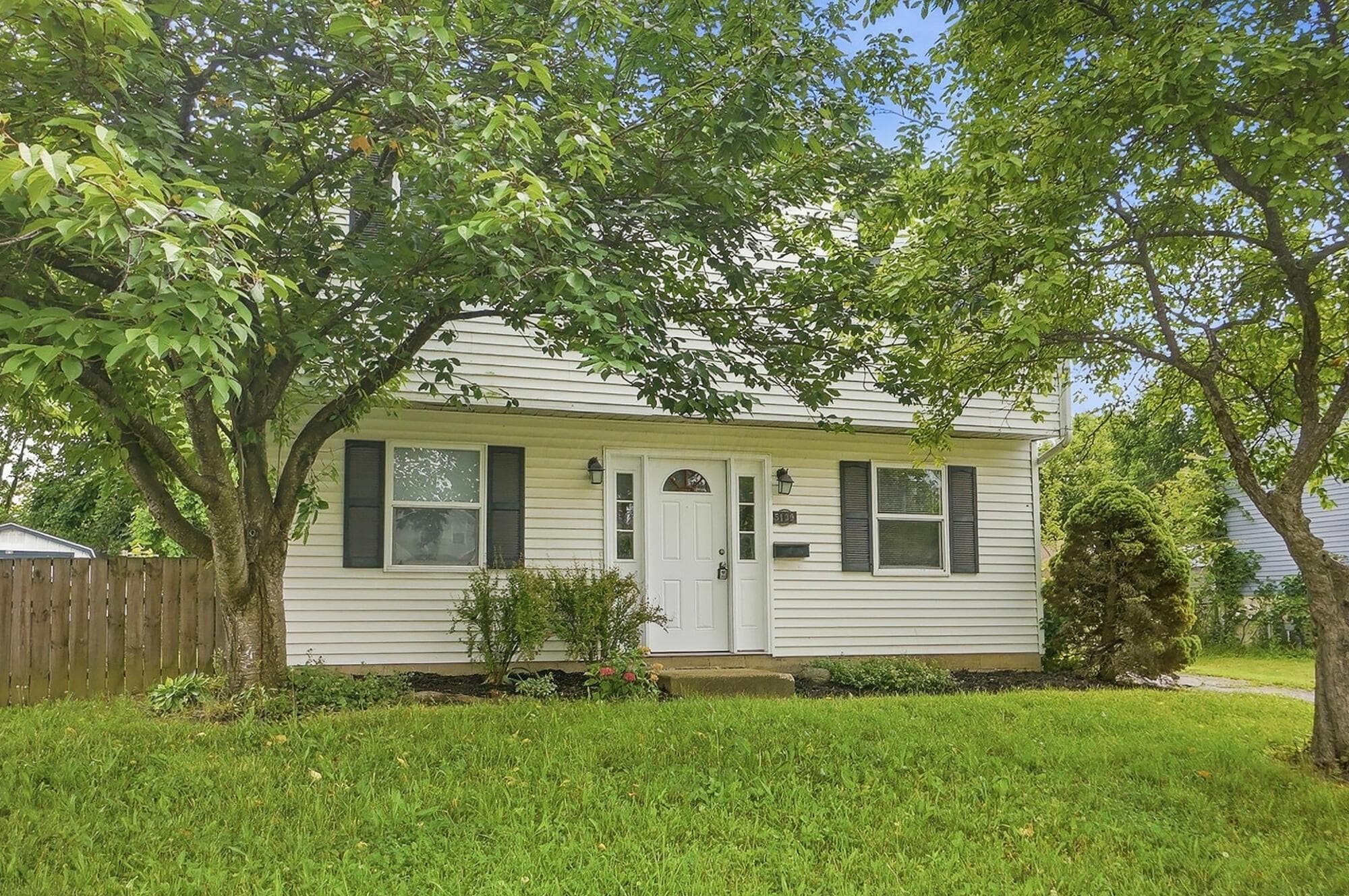 White siding exterior with dark shutters, prominent mature trees, green lawn, and a light-colored entry door