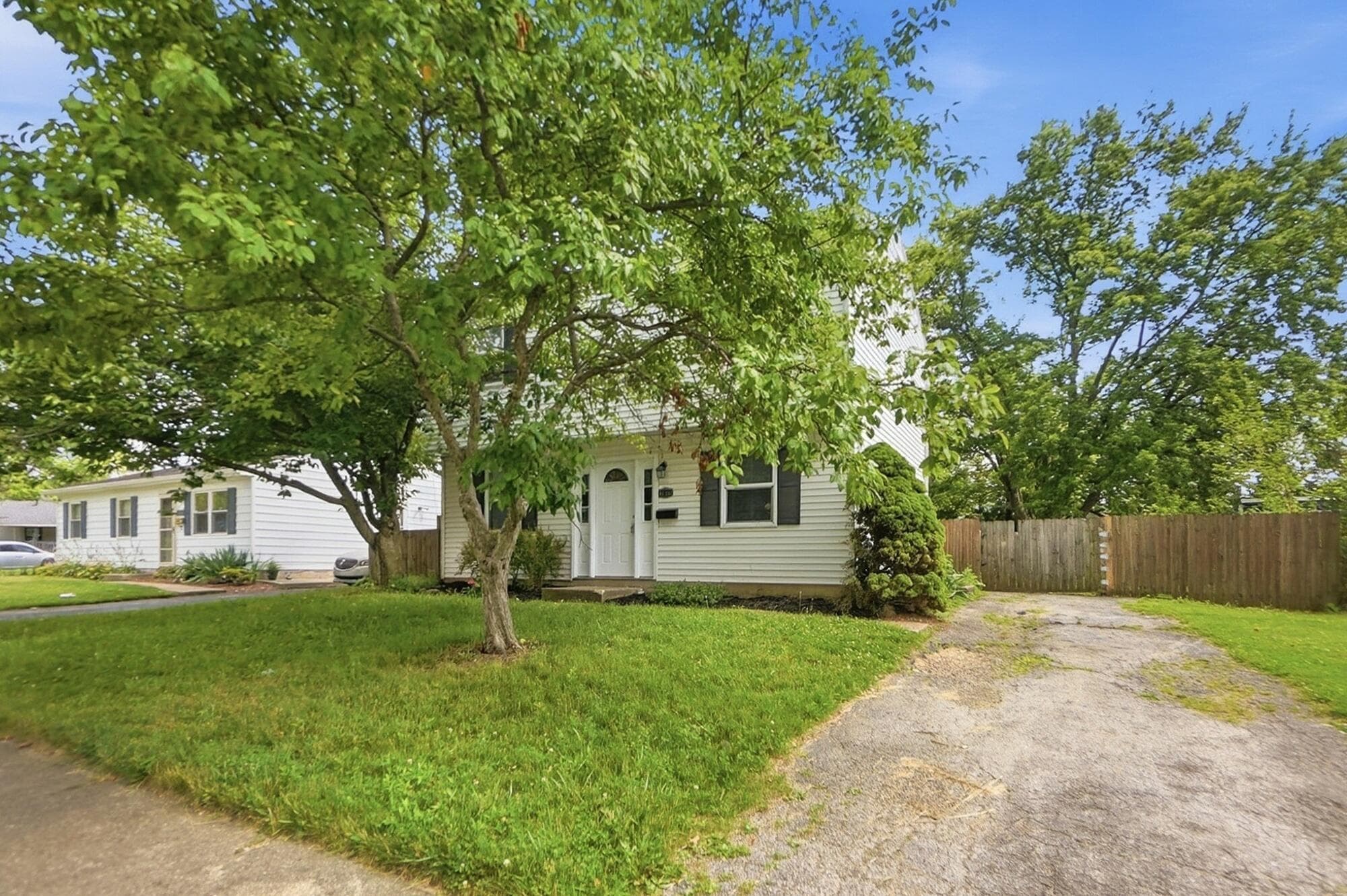 White siding exterior with dark shutters, established front lawn, mature trees, and a private driveway