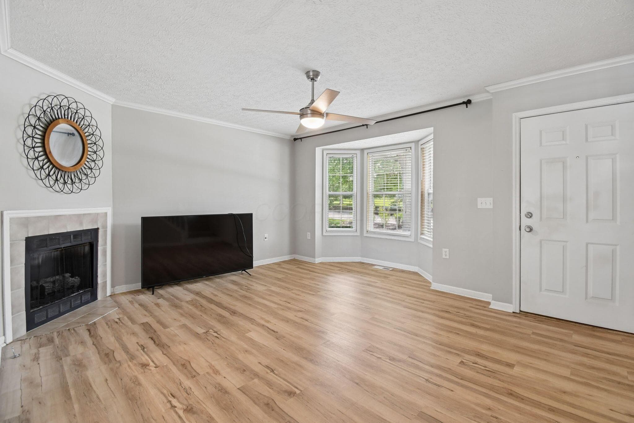 Wood-finish flooring throughout, bay windows with blinds, corner fireplace with tile surround, ceiling fan with integrated lighting, and a white paneled door