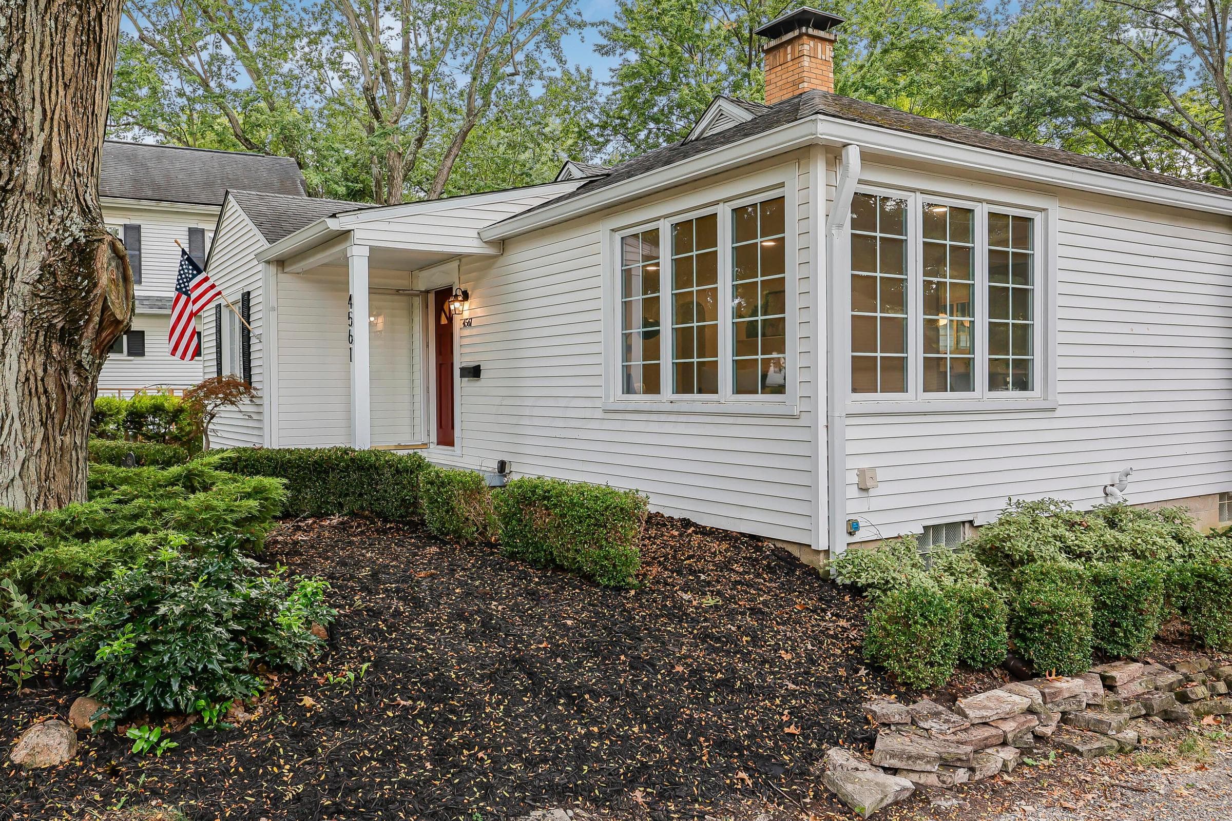 White siding exterior with a prominent bay window, covered entry, brick chimney, and mature landscaping