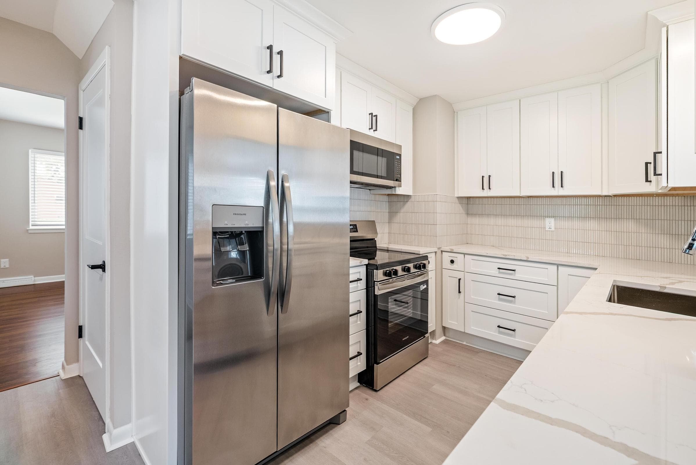 Stainless steel appliances, white cabinetry with black hardware, light-toned wood-finish flooring, white countertops, and a textured tile backsplash