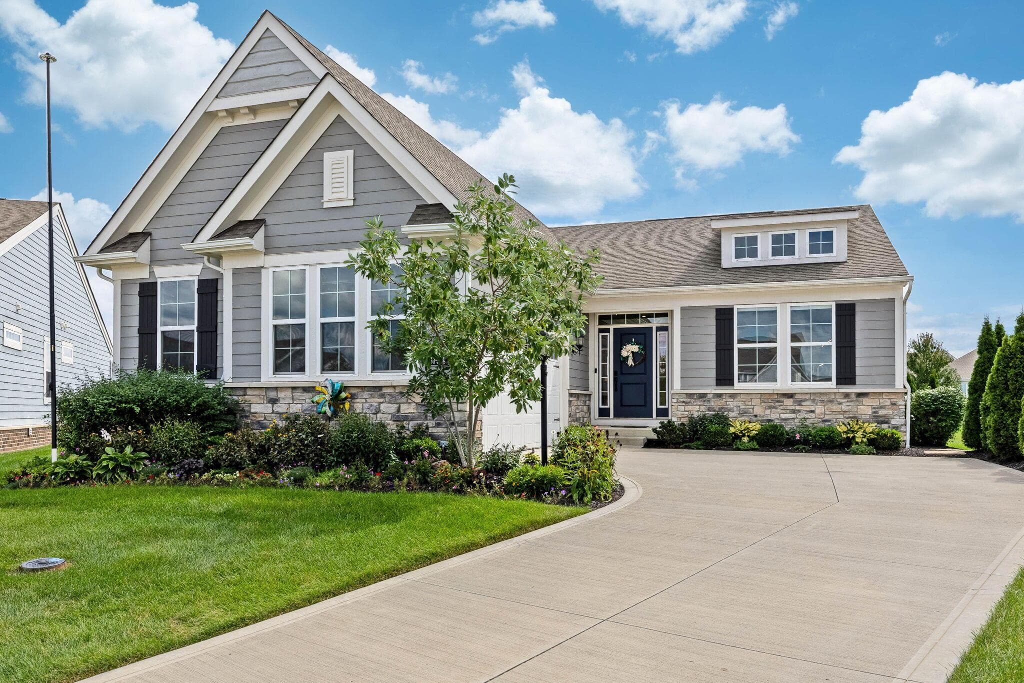 Grey siding exterior with stone veneer accents, a dark blue front door, dormer window, and an expansive concrete driveway