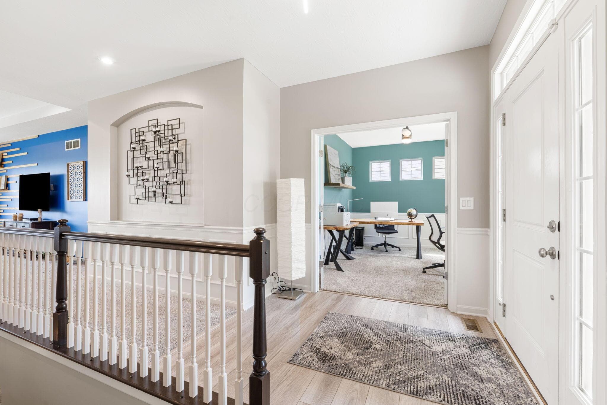 Wood-finish flooring, white paneled door with transom window, staircase with dark wood handrail and white spindles, architectural wall niche, and an open doorway to a carpeted room with teal walls