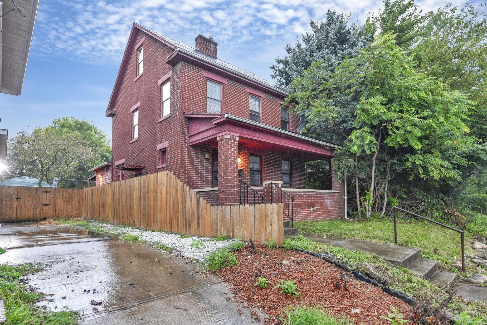 Brick exterior with a covered front porch, wood fencing, concrete driveway, and mature landscaping