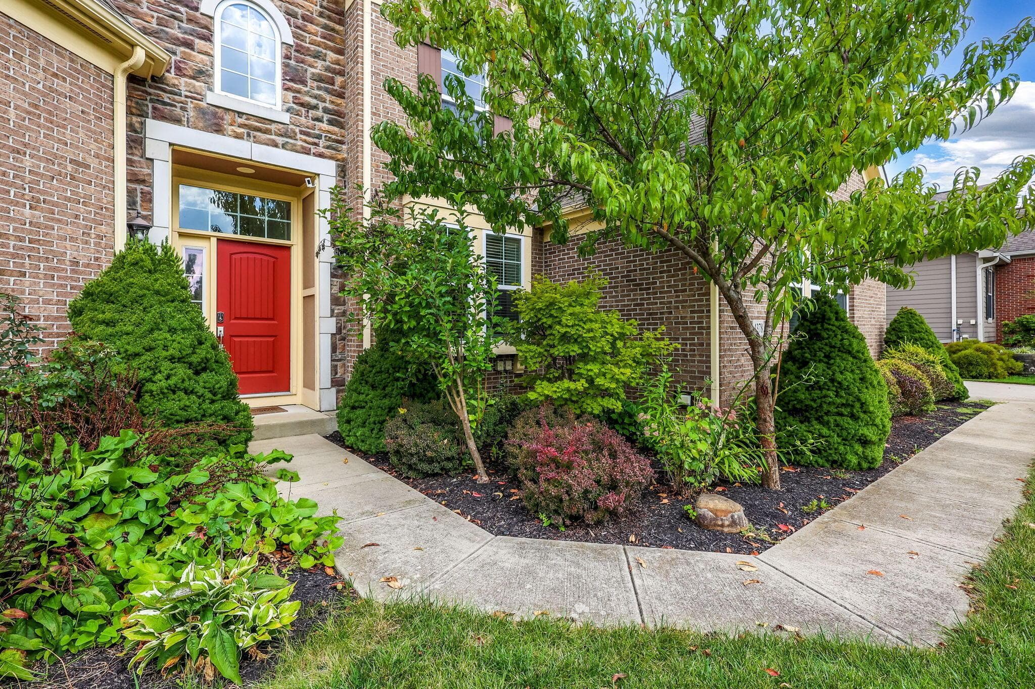 Brick exterior with stone accents, a vibrant red entry door with transom and sidelights, and mature front landscaping