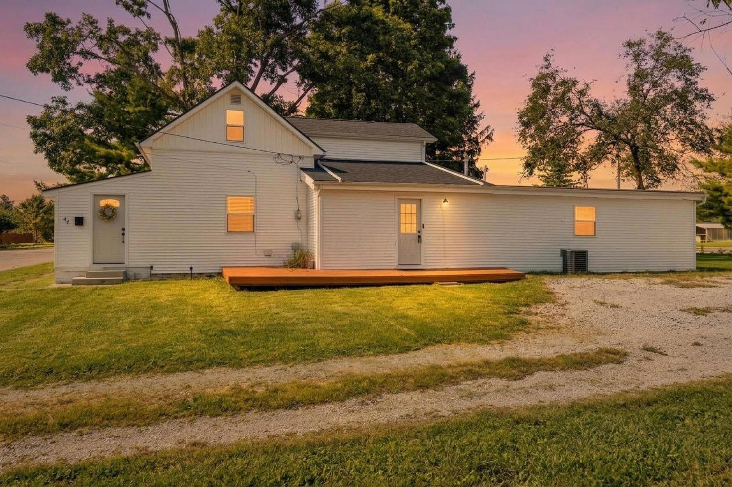 White siding exterior with a dark shingle roof, a ground-level deck, and a gravel driveway