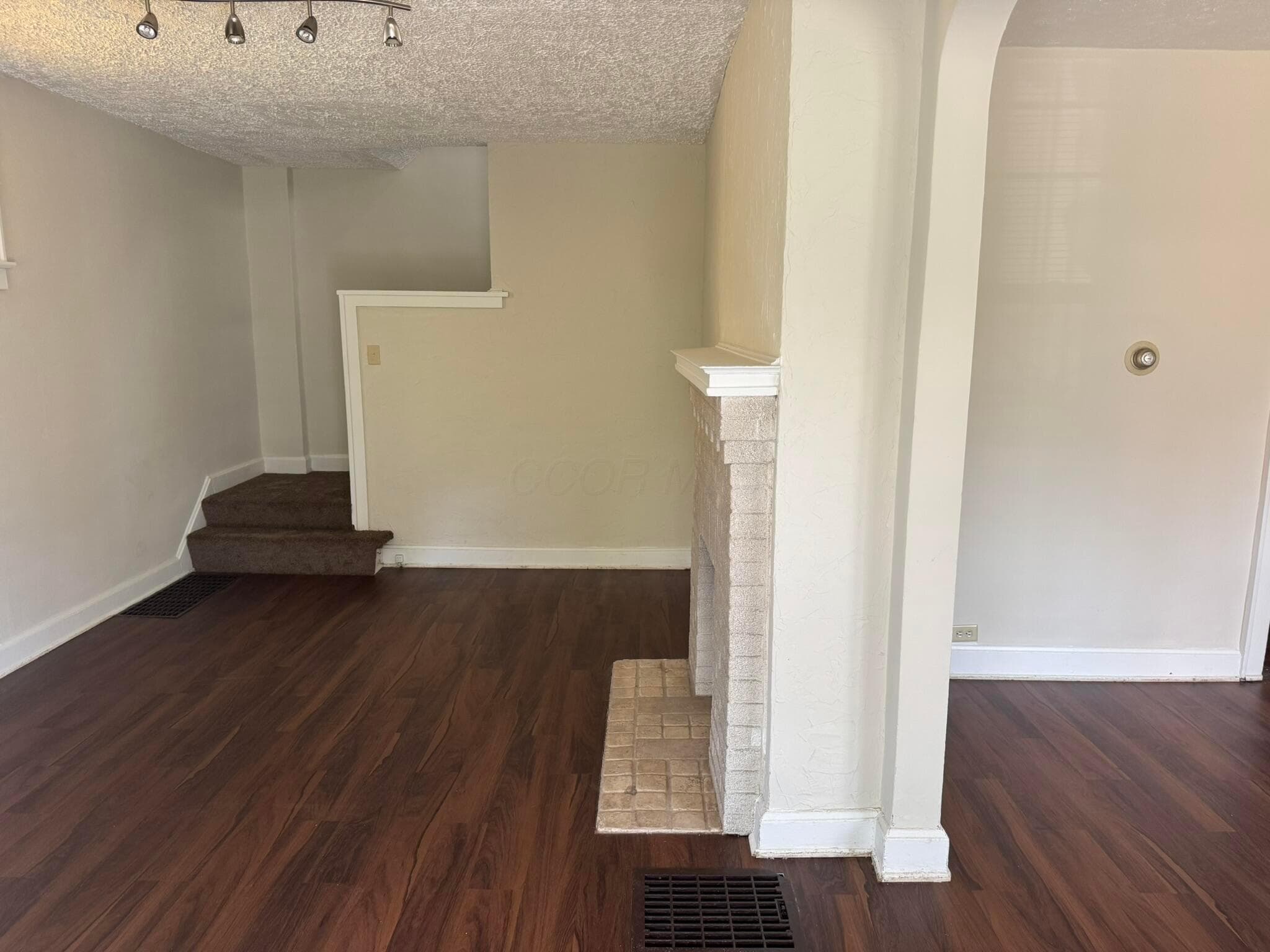 Wood-finish flooring throughout, featuring a white brick fireplace with a mantel, and a track lighting fixture