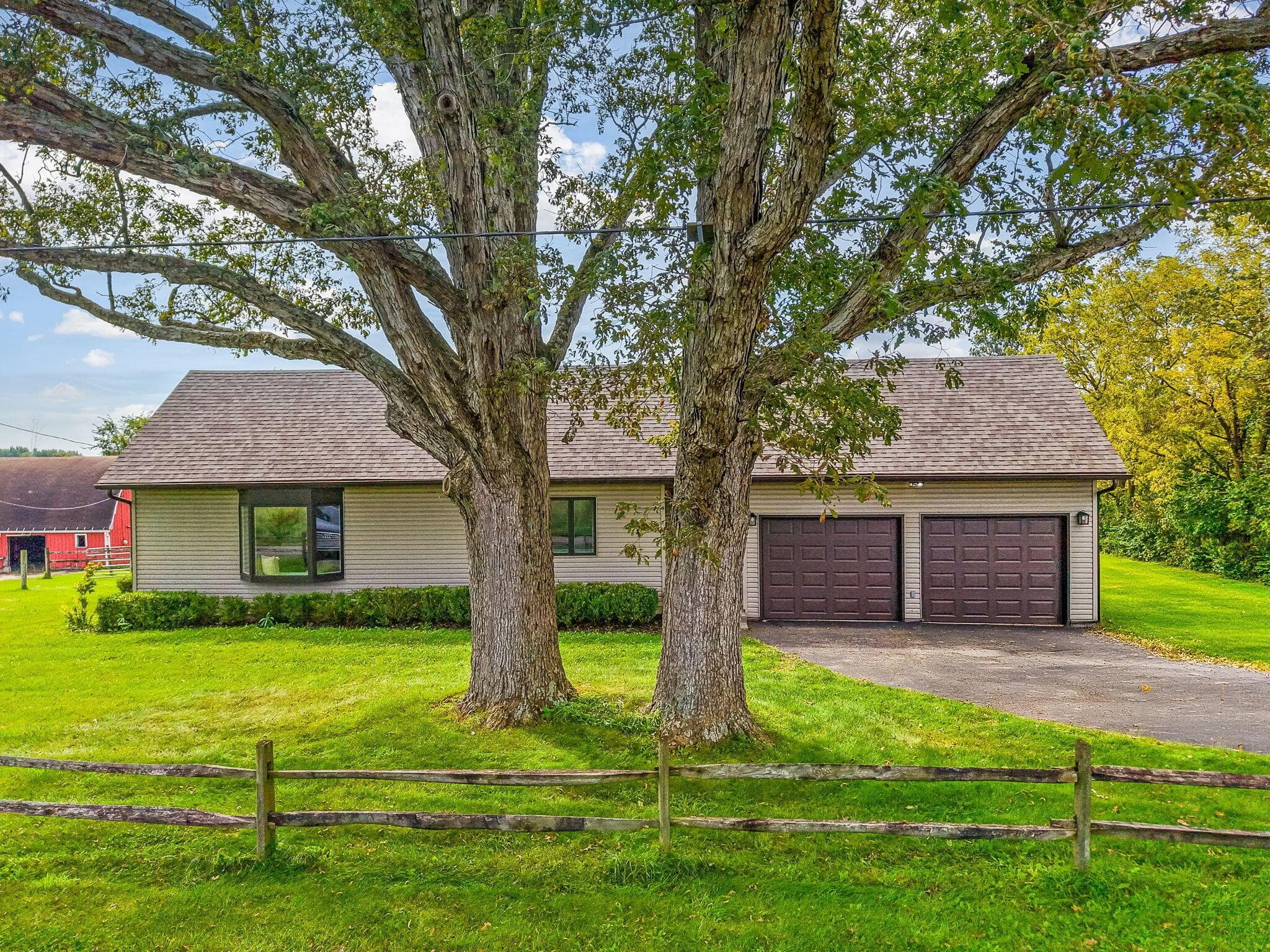Mature trees framing the property, a two-car garage with panel doors, and a wood split-rail fence
