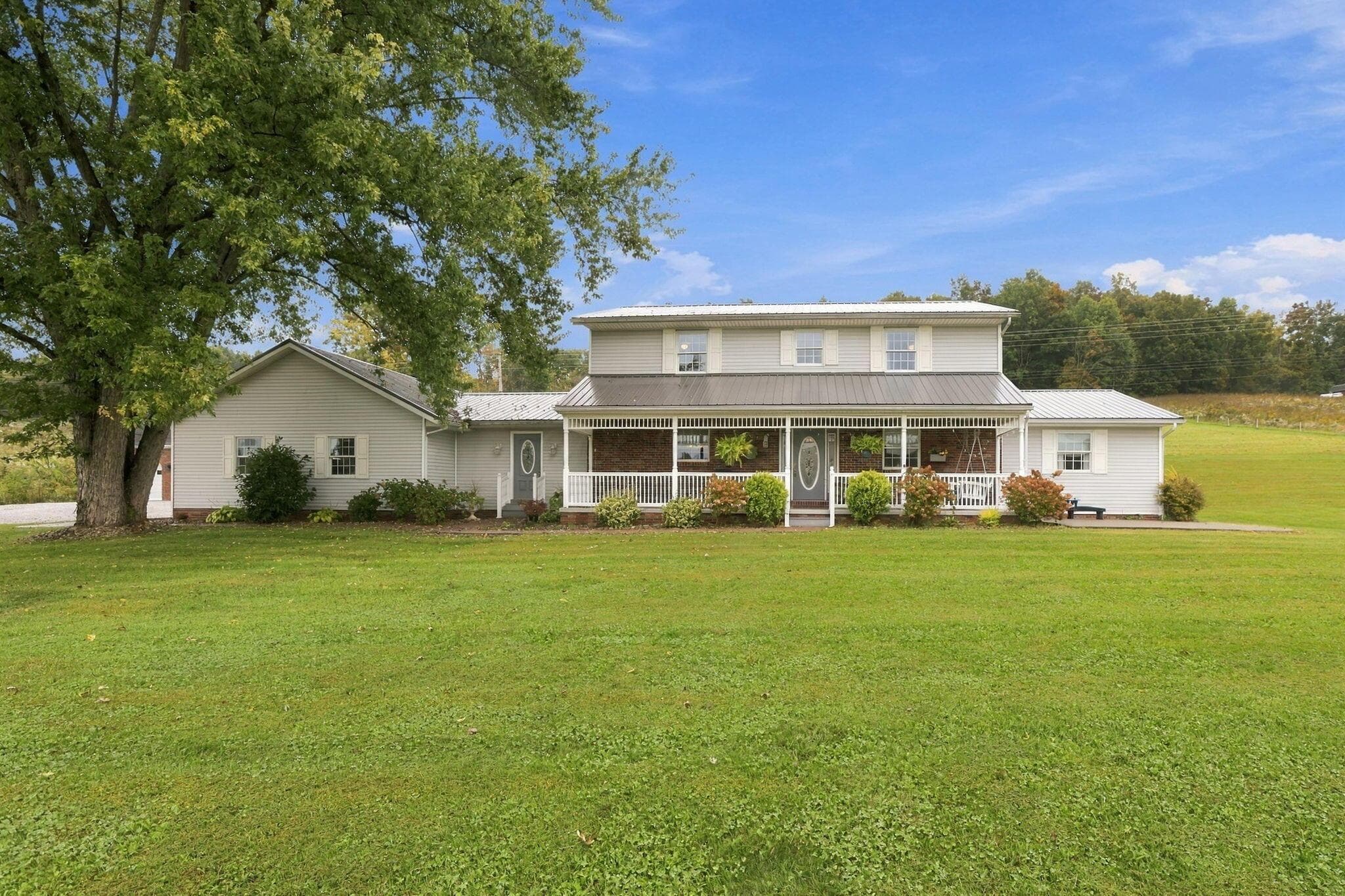 Expansive green lawn surrounds a two-story home with a wrap-around porch, metal roof, and light-toned siding