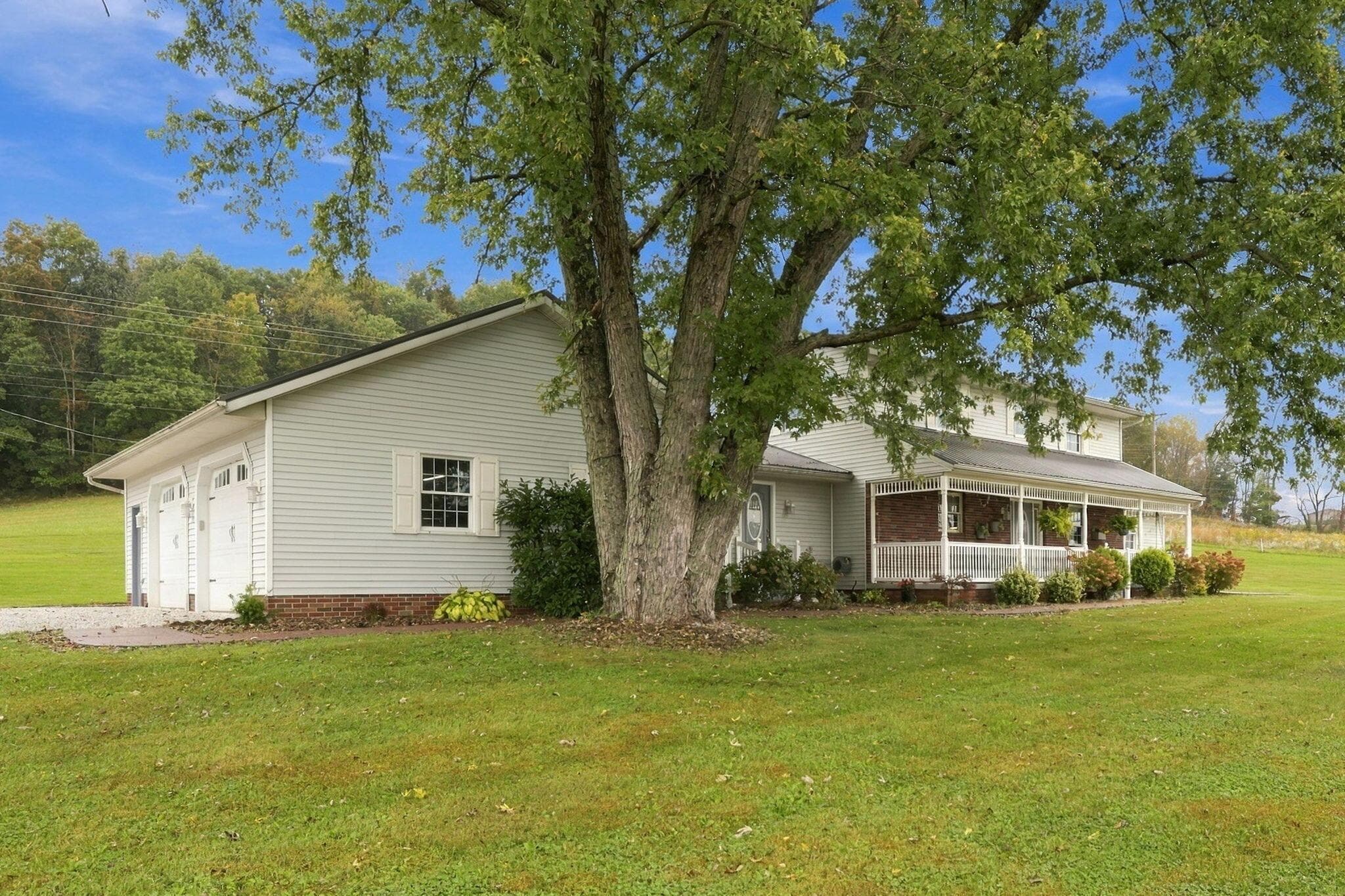 Expansive green lawn surrounds the residence, featuring a covered porch with white railings, a multi-car garage, and mature landscaping