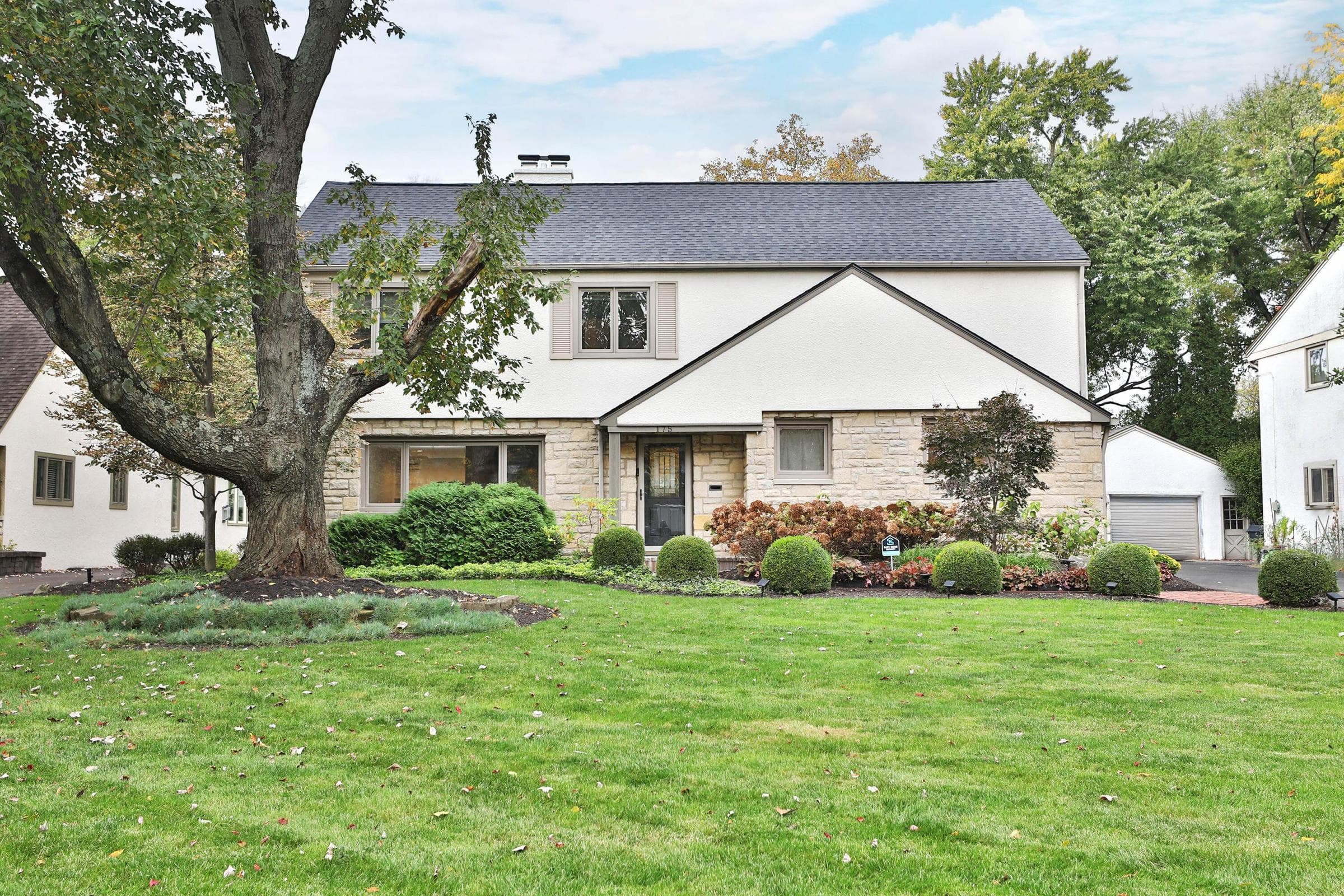Two-story residence featuring stone and stucco exterior, shingle roof, chimney stack, and mature landscaping