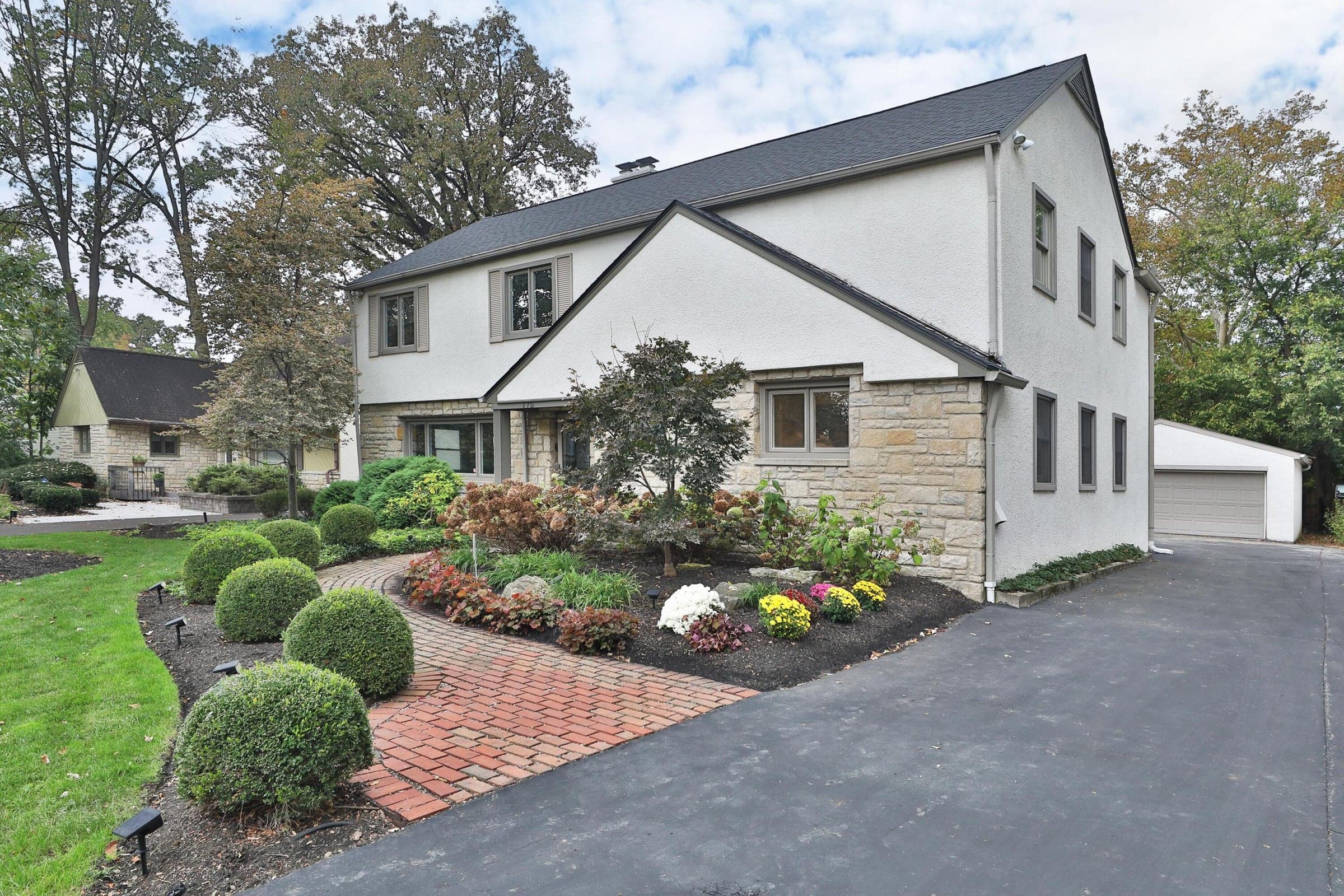 Exterior facade featuring a combination of stone masonry and stucco, multi-level gabled roofline, brick paver walkway, mature landscape beds, and an asphalt driveway
