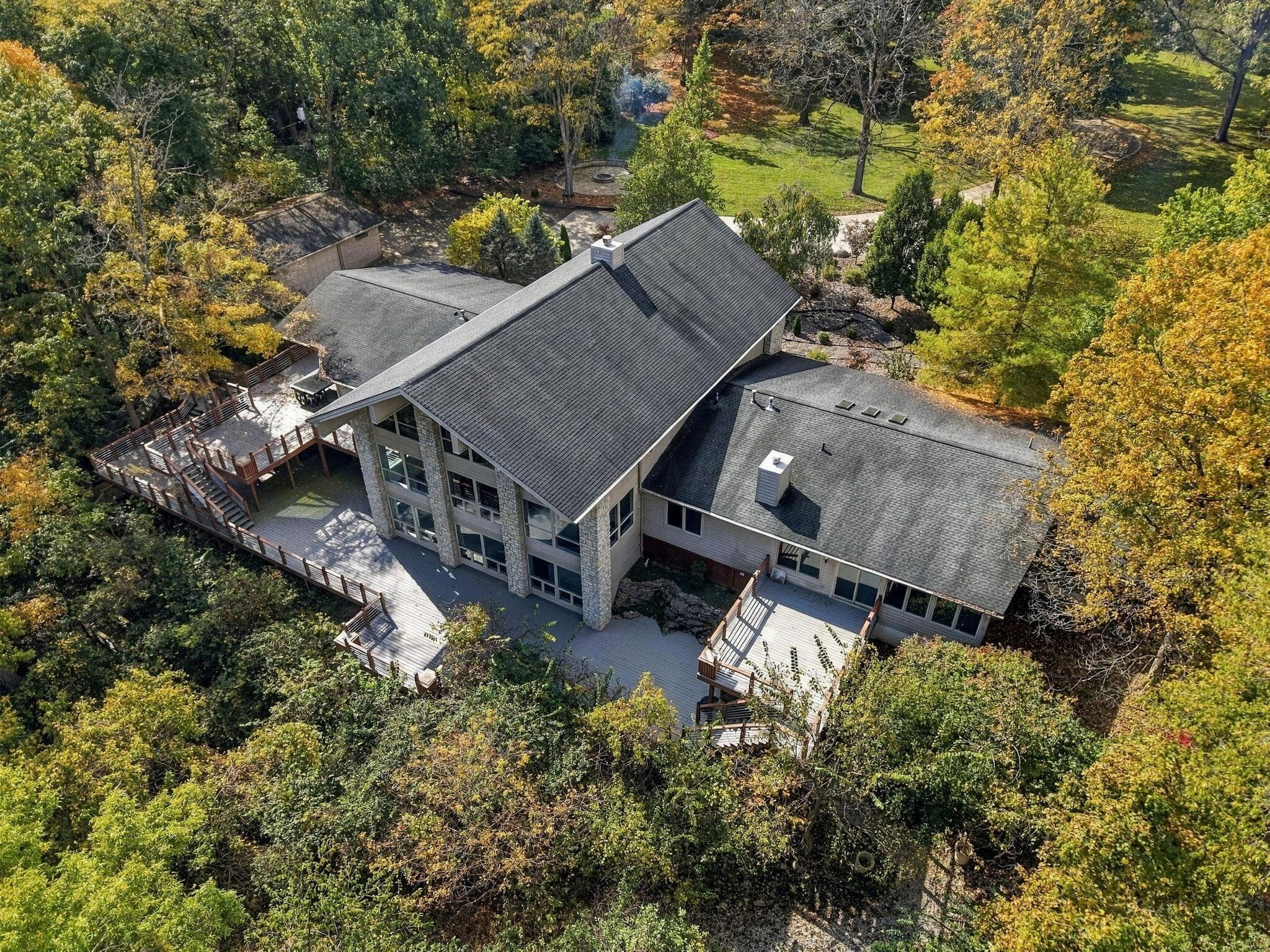 Multi-level residence featuring stone siding, expansive multi-tier wooden decking, two prominent chimneys, and shingle roofing, all situated within a wooded lot