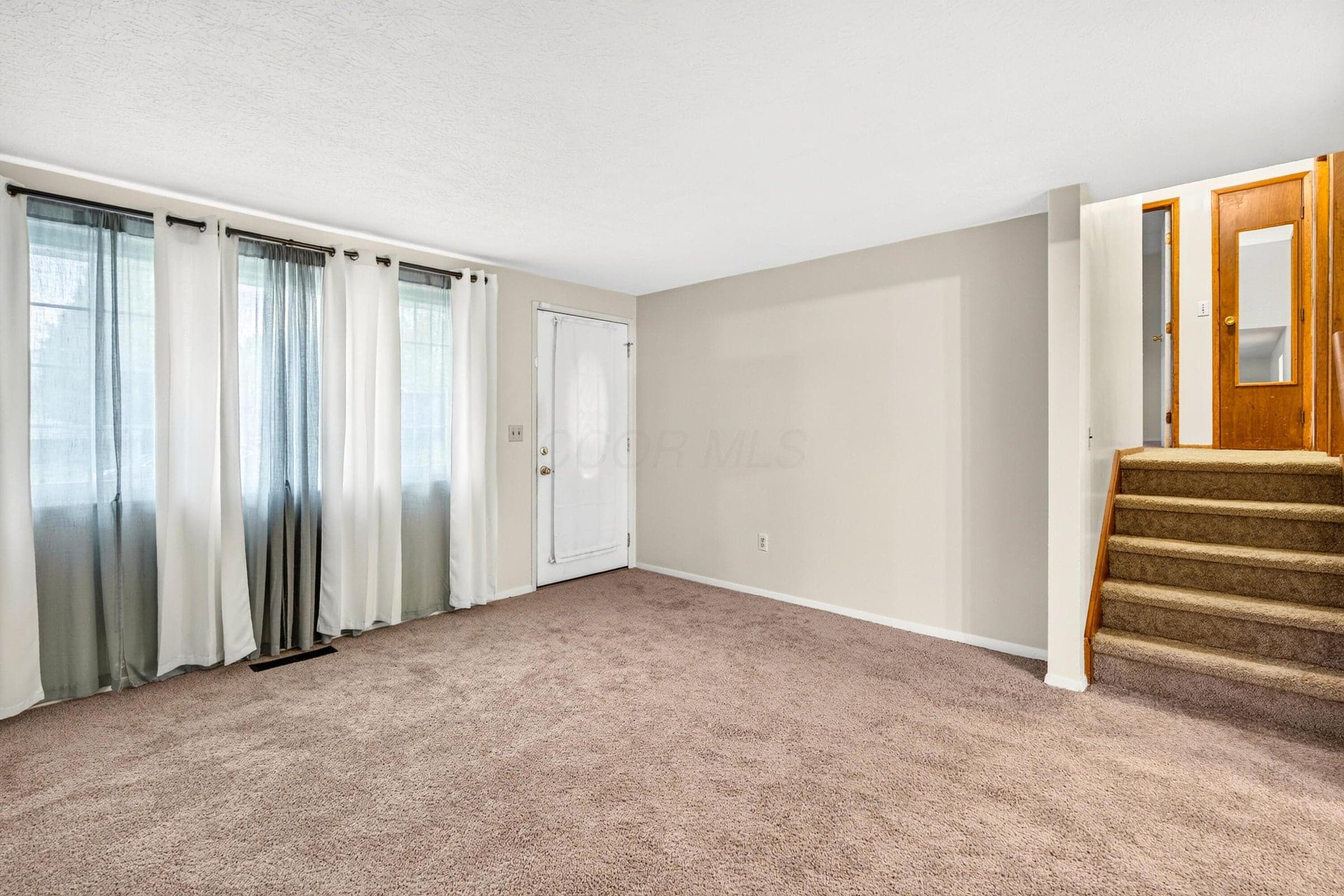 Neutral interior space featuring carpeted flooring, a bank of large windows with natural light, and a wooden staircase