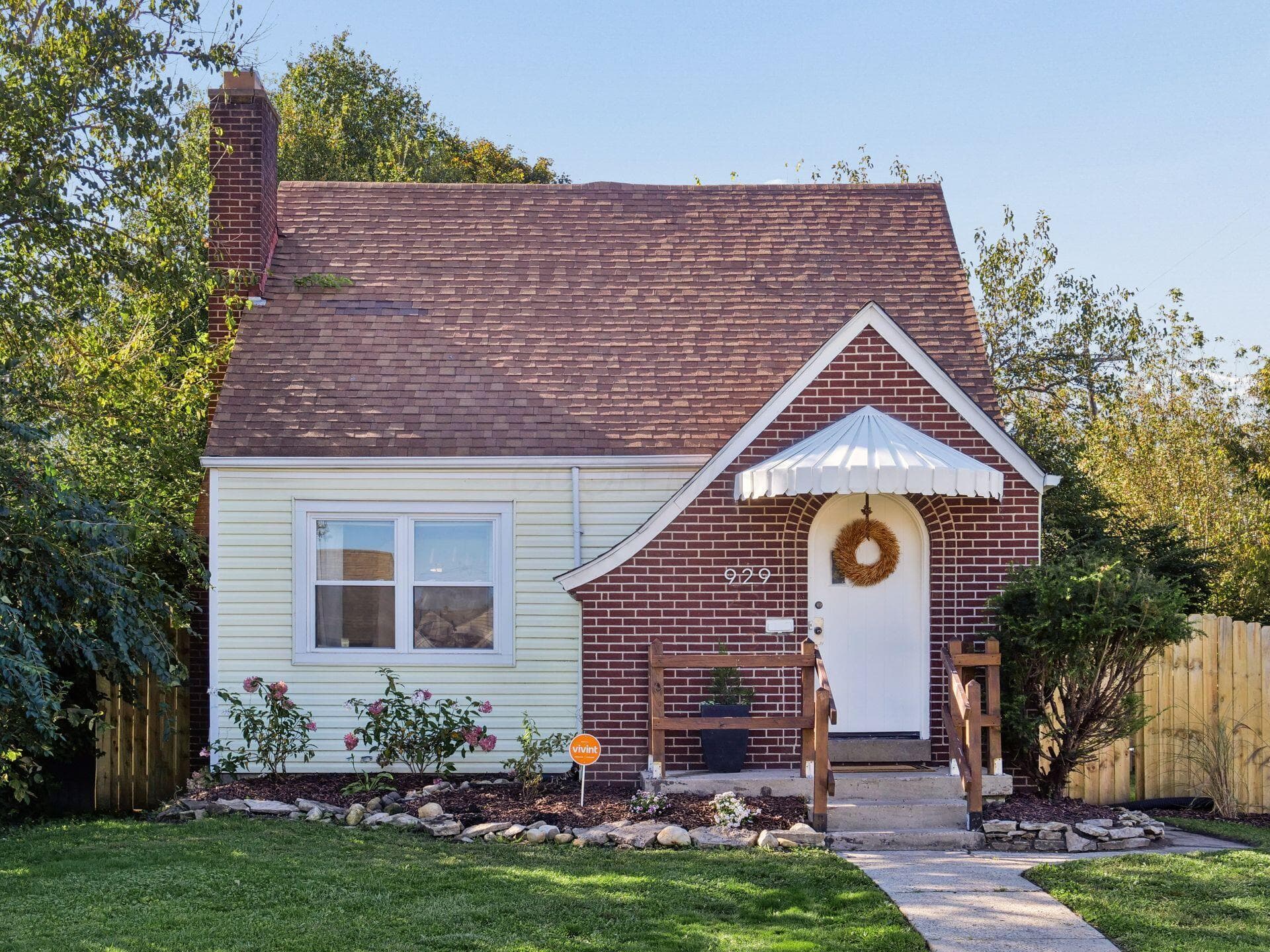 Exterior featuring red brick and light-toned siding, a pitched roofline, a decorative arched entryway with a scalloped metal awning, a prominent brick chimney, and stone-bordered landscaping
