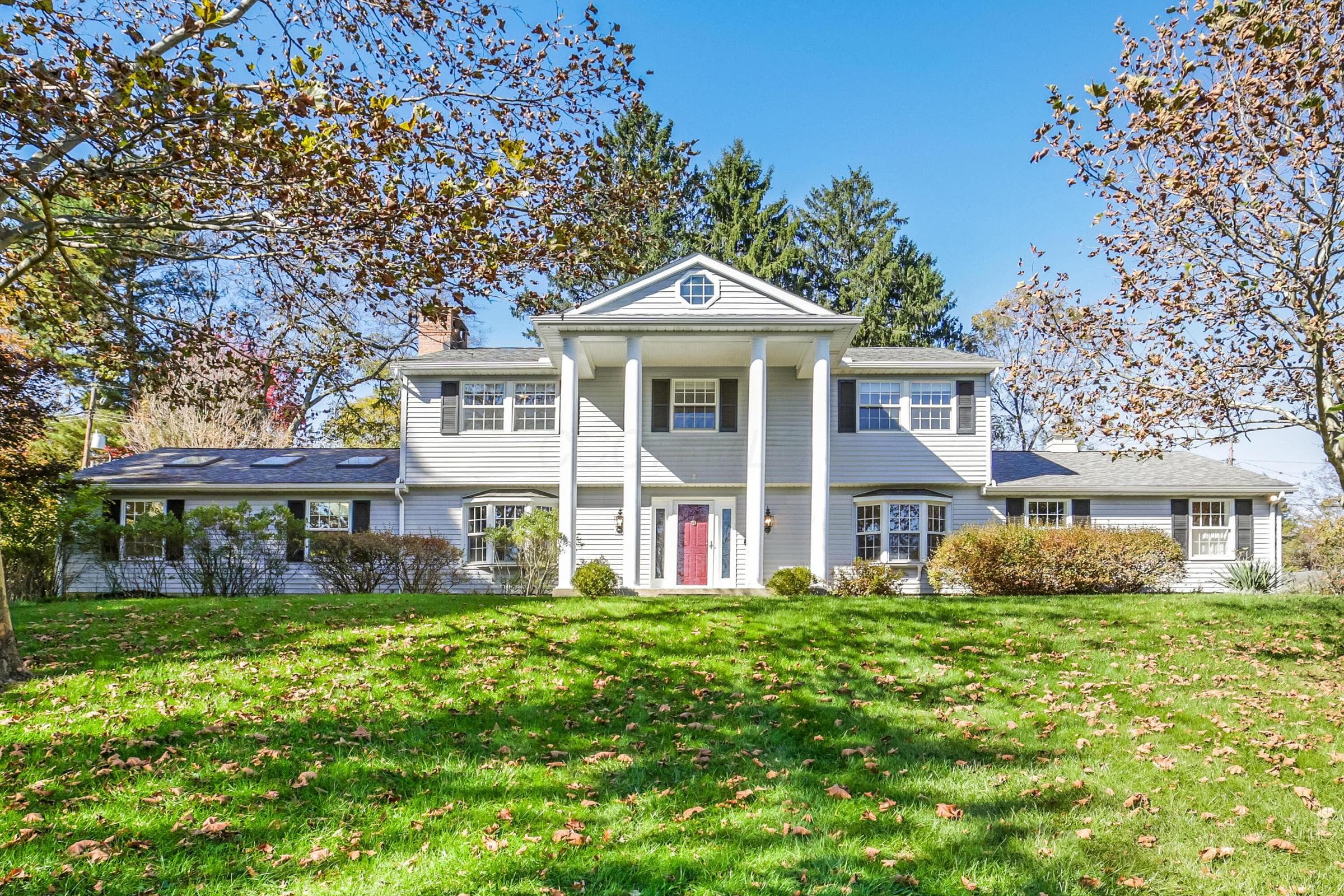 Colonial-style exterior featuring a covered front porch with prominent columns, a central pediment, bay windows, and skylights set against a mature landscape