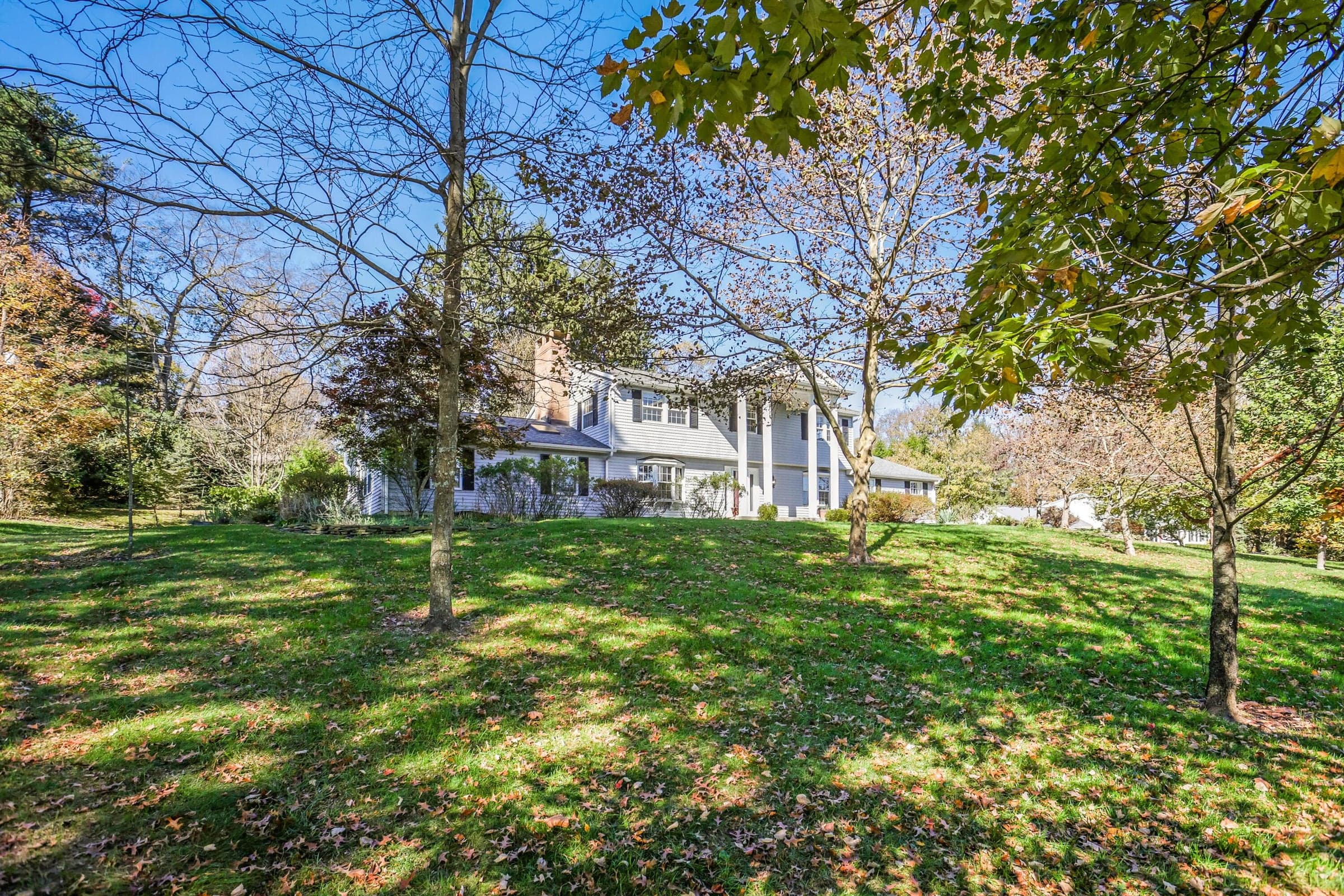 Two-story Colonial exterior featuring a covered porch with tall columns, light-colored siding, dark shutters, and a brick chimney, situated on a spacious, tree-lined lot