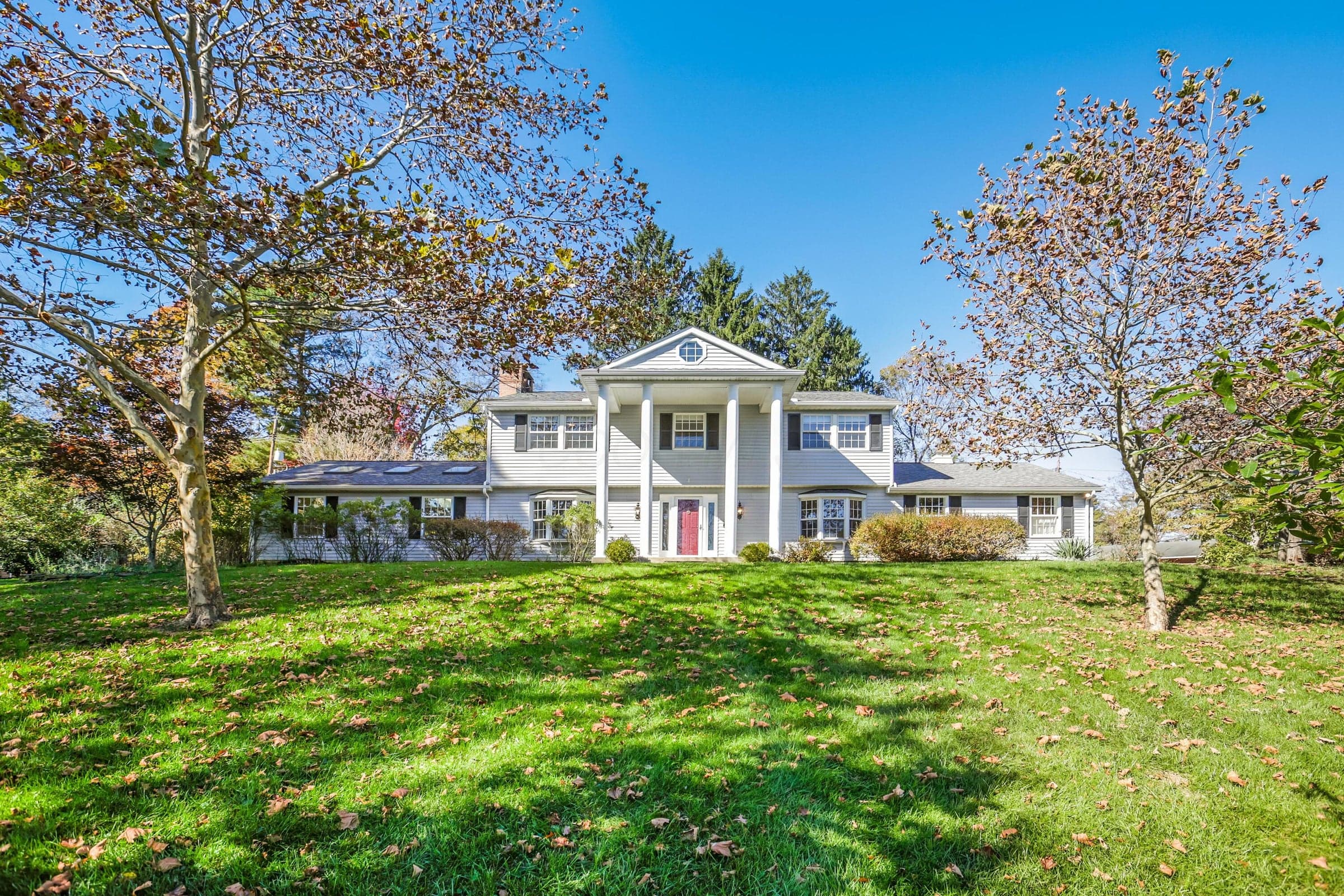 Colonial-style exterior featuring a prominent two-story portico with classical columns, a centered pediment with an oculus window, symmetrical window shutters, and skylights set against an expansive lawn