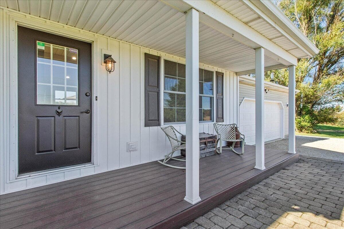 Covered front porch featuring wood-finish decking, vertical siding, shutter-framed windows, and an exterior wall-mounted light fixture