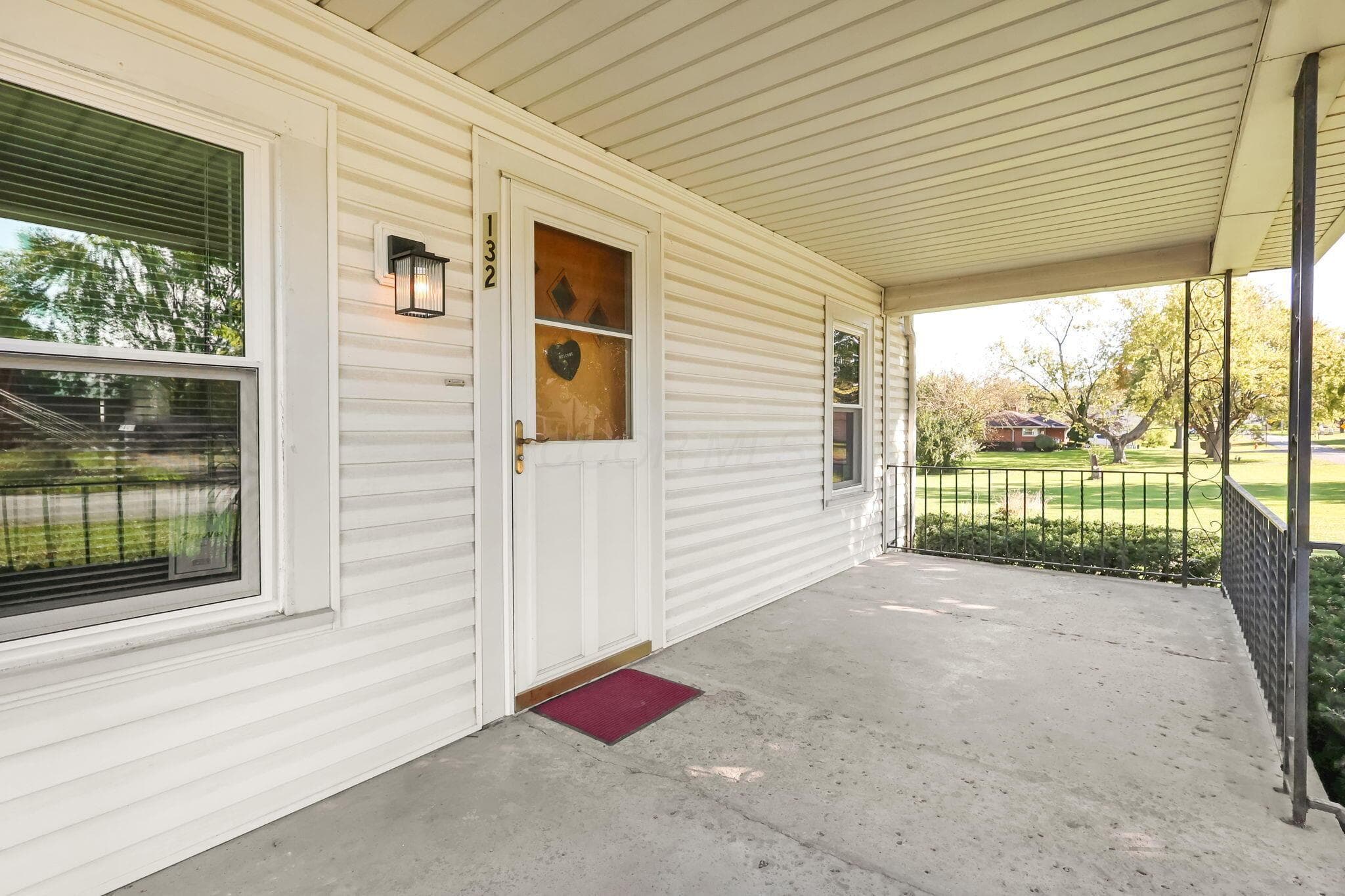 Covered exterior porch featuring concrete flooring, horizontal siding, metal railings, and a glass-panel entry door with an attached wall-mounted light fixture