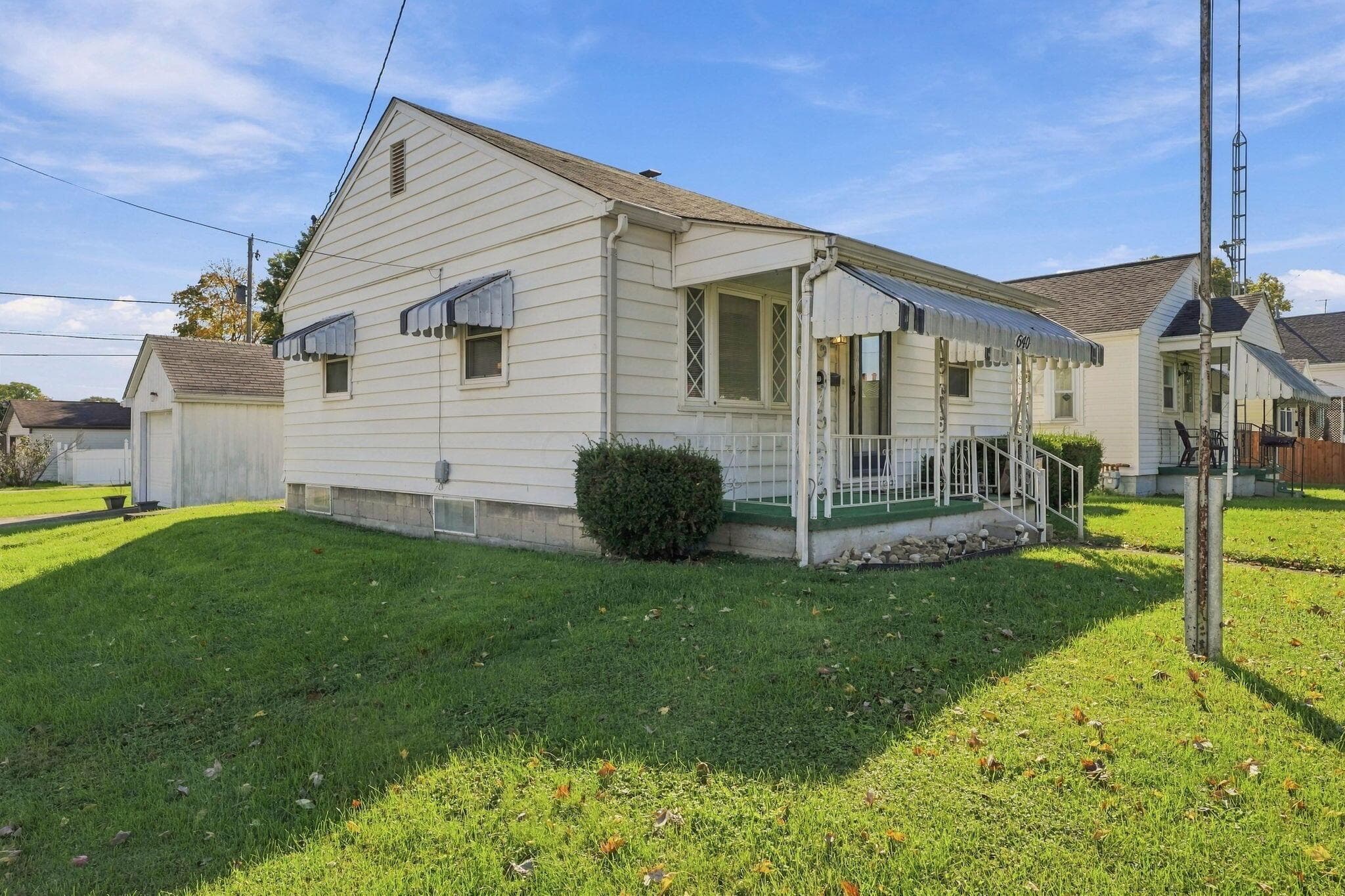 Single-story residence featuring light-colored horizontal siding, a front porch with metal railings, metal window awnings, and a detached garage